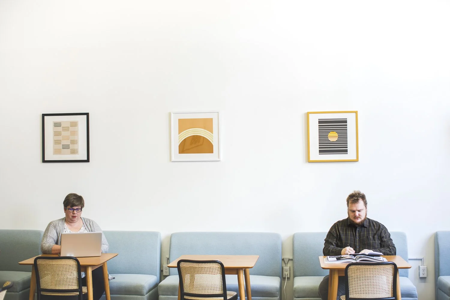 A cozy, vibrant space showcases a white woman typing on a laptop and a bearded man reading, surrounded by warm artwork, embodying fat joy, creativity, and community connection.