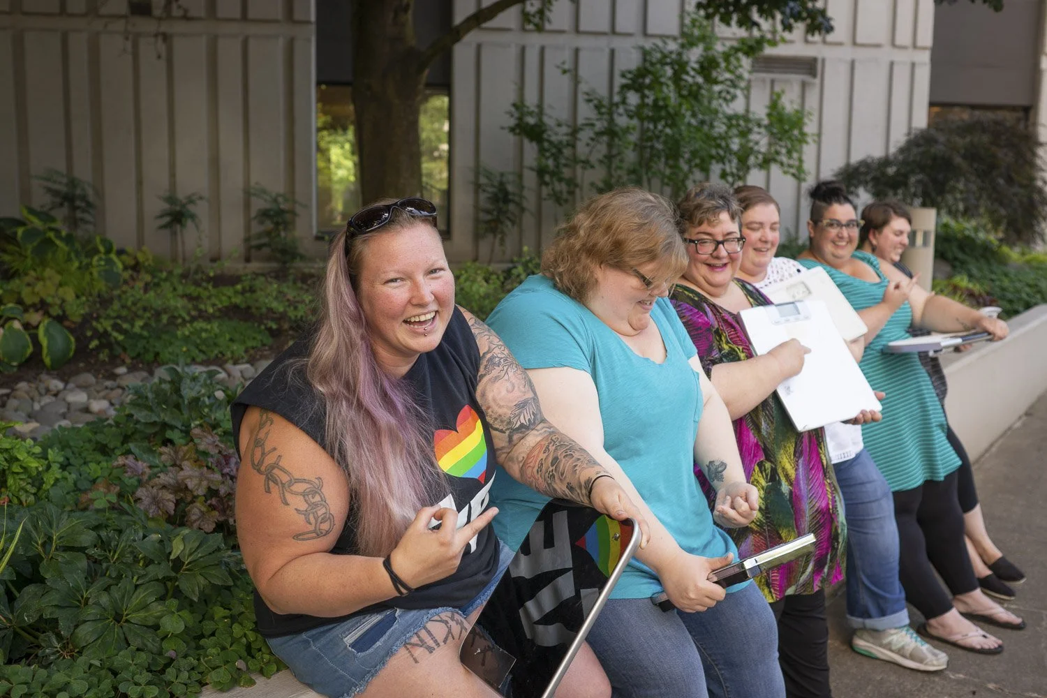 A joyful group of diverse plus-size women, showcasing vibrant tattoos and colorful clothing, laughing together in a lush, green setting, embodying fat liberation and body positivity in an inclusive moment.