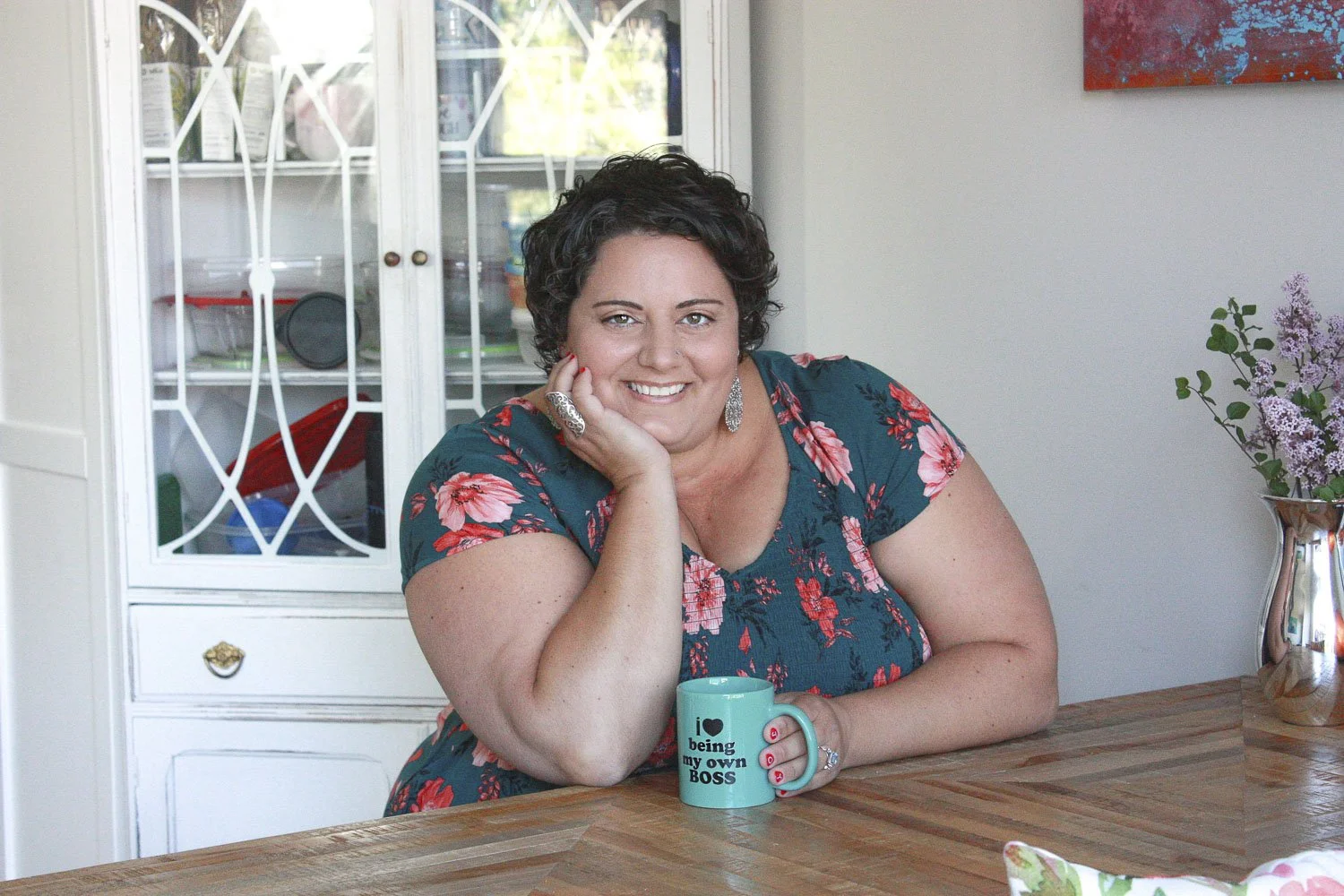 A woman with short dark curly hair, wearing a floral dress, smiling and resting her face on her left hand while sitting at a wooden table. She has a tattoo on her right wrist and large earrings. In front of her is a coffee mug that reads "I love being my own boss."