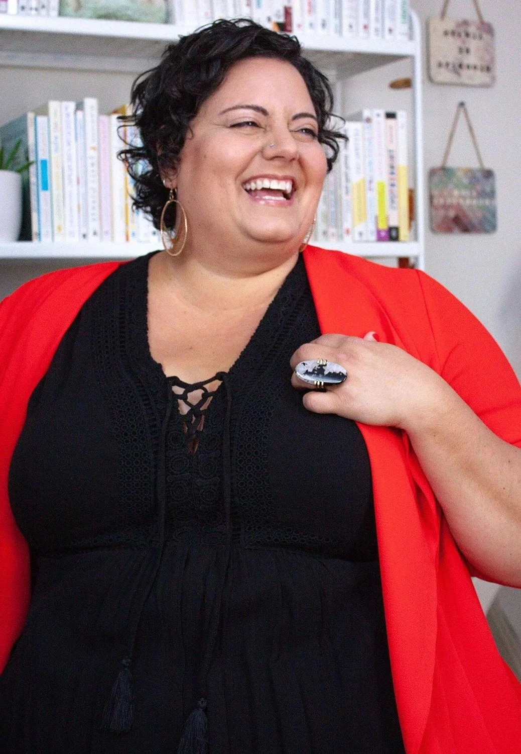 Sophia Apostol,  the founder of Fat Joy Creation Lab, and a joyful, plus-size white woman with curly brown hair, beams in a vibrant red jacket over a black dress, radiating body positivity and empowerment against a backdrop of colorful bookshelves.