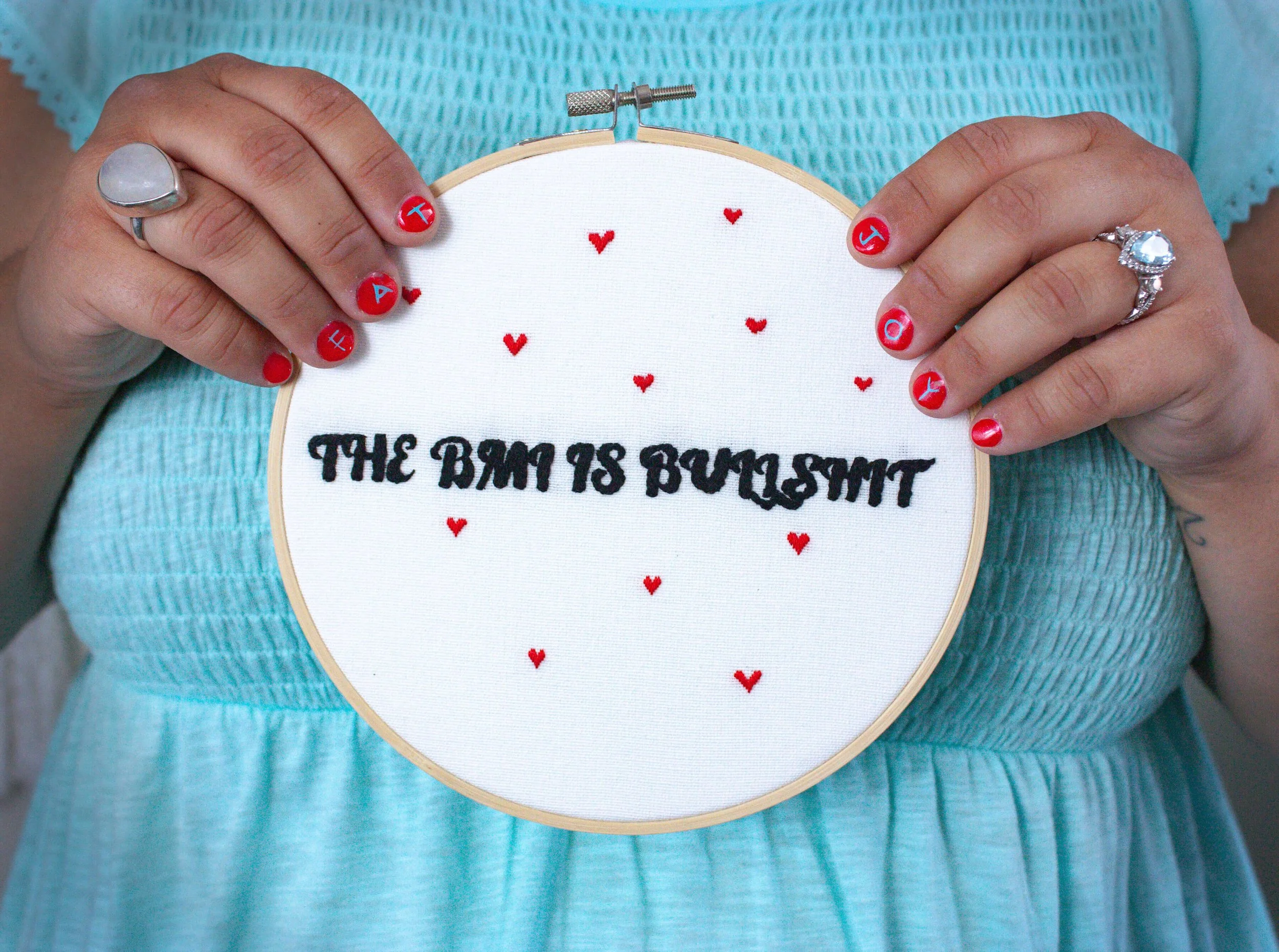 Close-up of hands holding an embroidery hoop with text and small red hearts, featuring finger nail art with letter and airplane symbols, and wearing multiple rings.