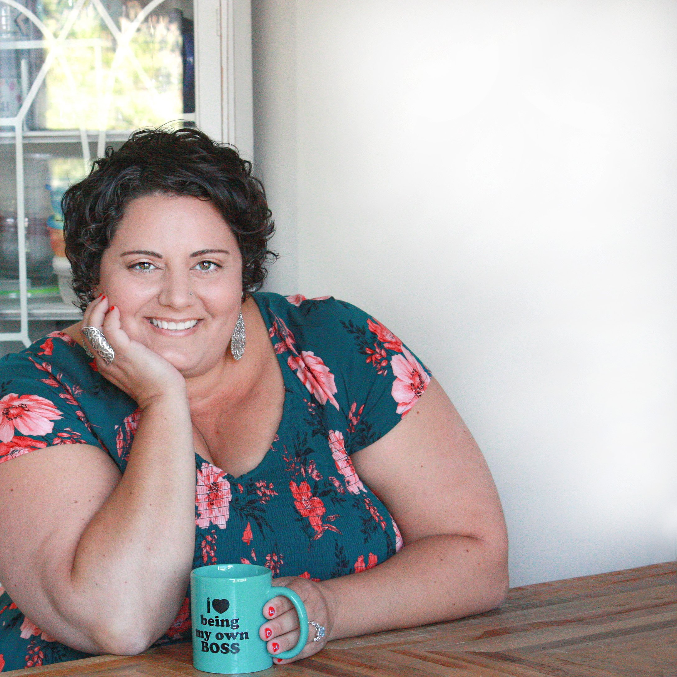 Sophia Apostol sitting at a table smiling at the camera holding a turquoise mug that reads I heart being my own boss.
