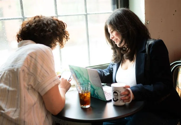 Two women sitting at a small round table near a window, looking at a laptop screen and smiling. One woman holds a mug, and there is a glass of iced tea on the table.