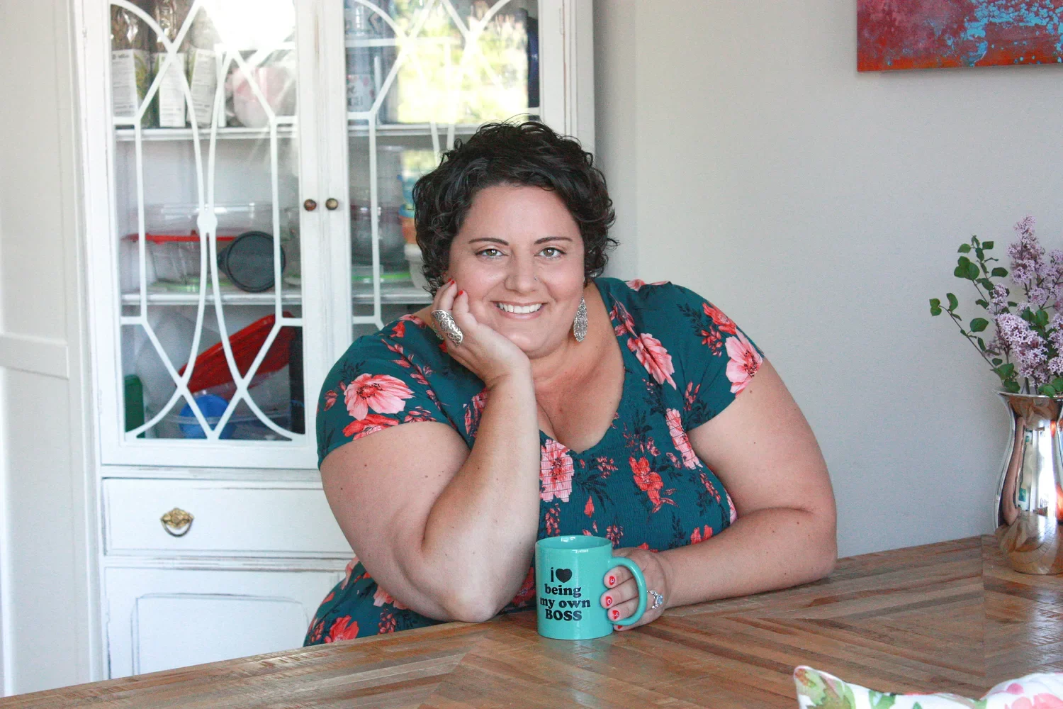 Body liberation coach smiling at a wooden table with a teal mug that reads 'I love being my own boss', wearing a floral dress