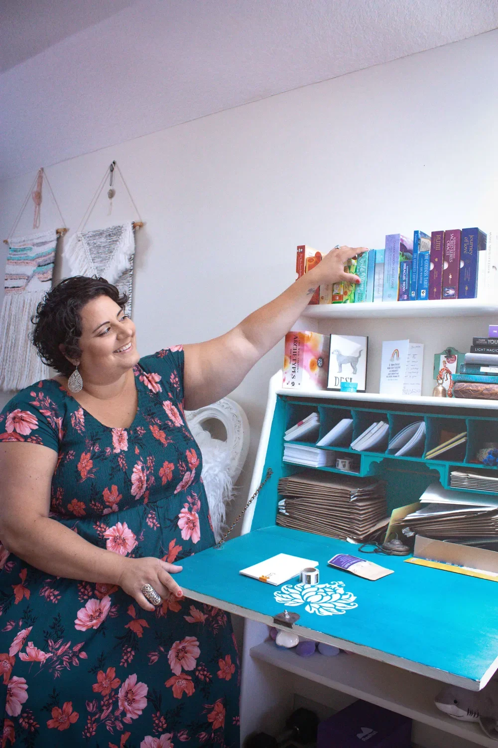 Fat Joy coach in a floral dress gesturing toward a rainbow-organized bookshelf above a painted teal secretary desk