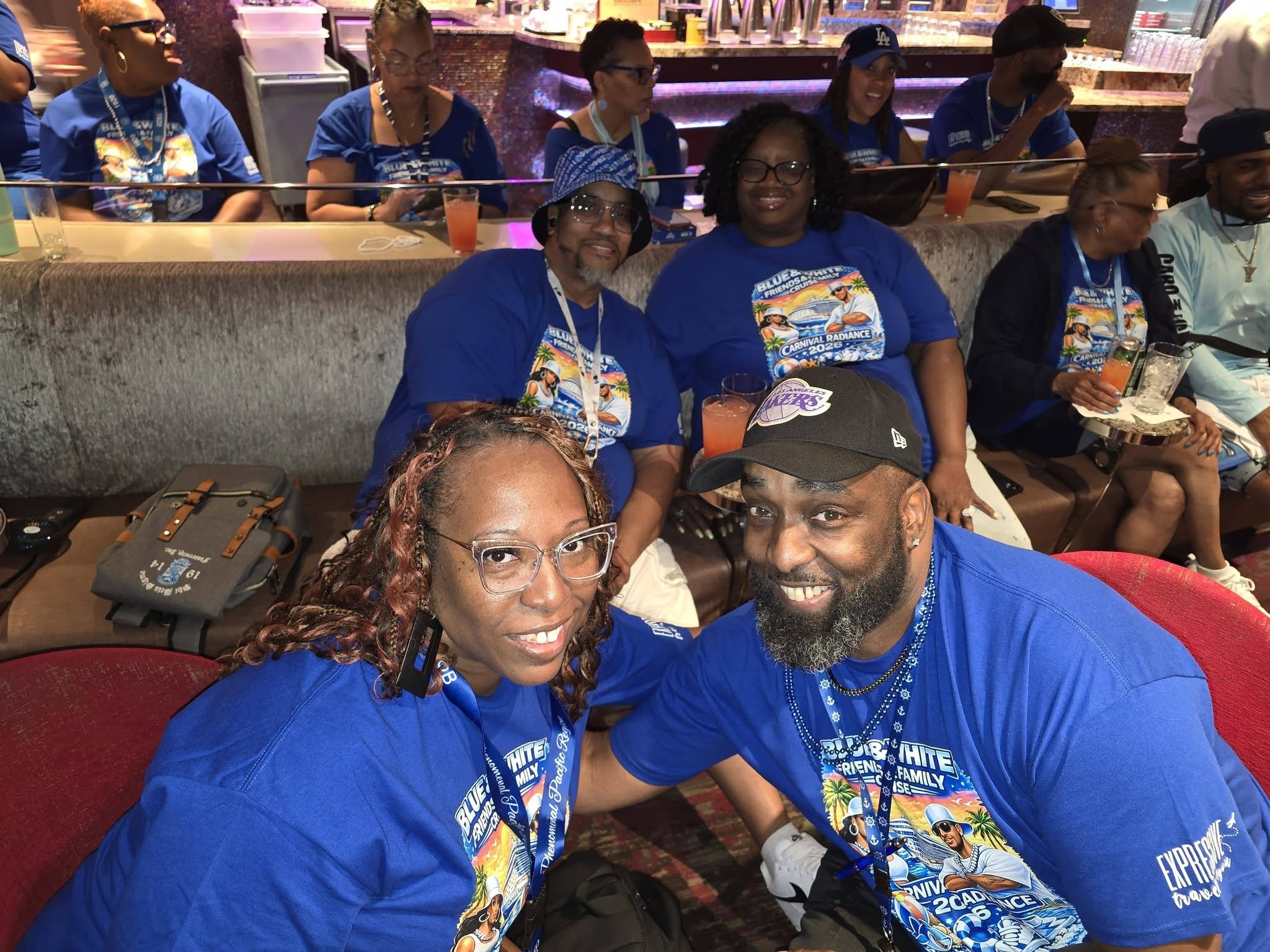 A group of people sitting together in a lounge area, wearing blue shirts and lanyards, attending a cruise event. Some are smiling and holding drinks, with a bar in the background.