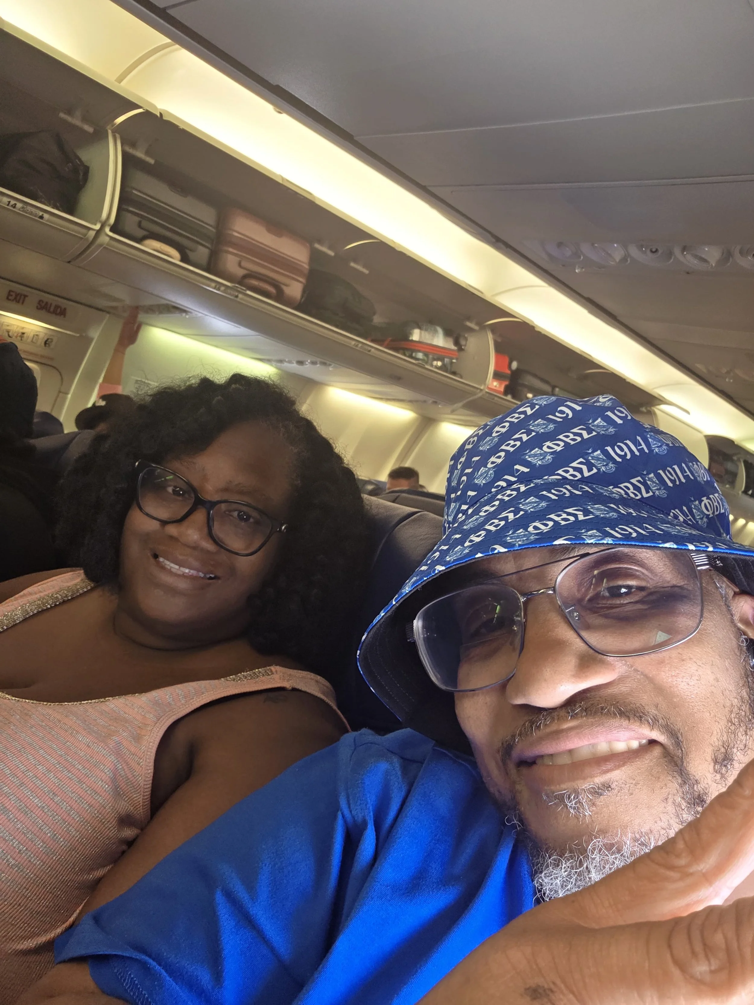 A selfie on an airplane showing a woman with glasses and curly hair, and a man wearing glasses and a blue cap with white writing, smiling at the camera. Overhead luggage compartments and airplane ceiling are visible.