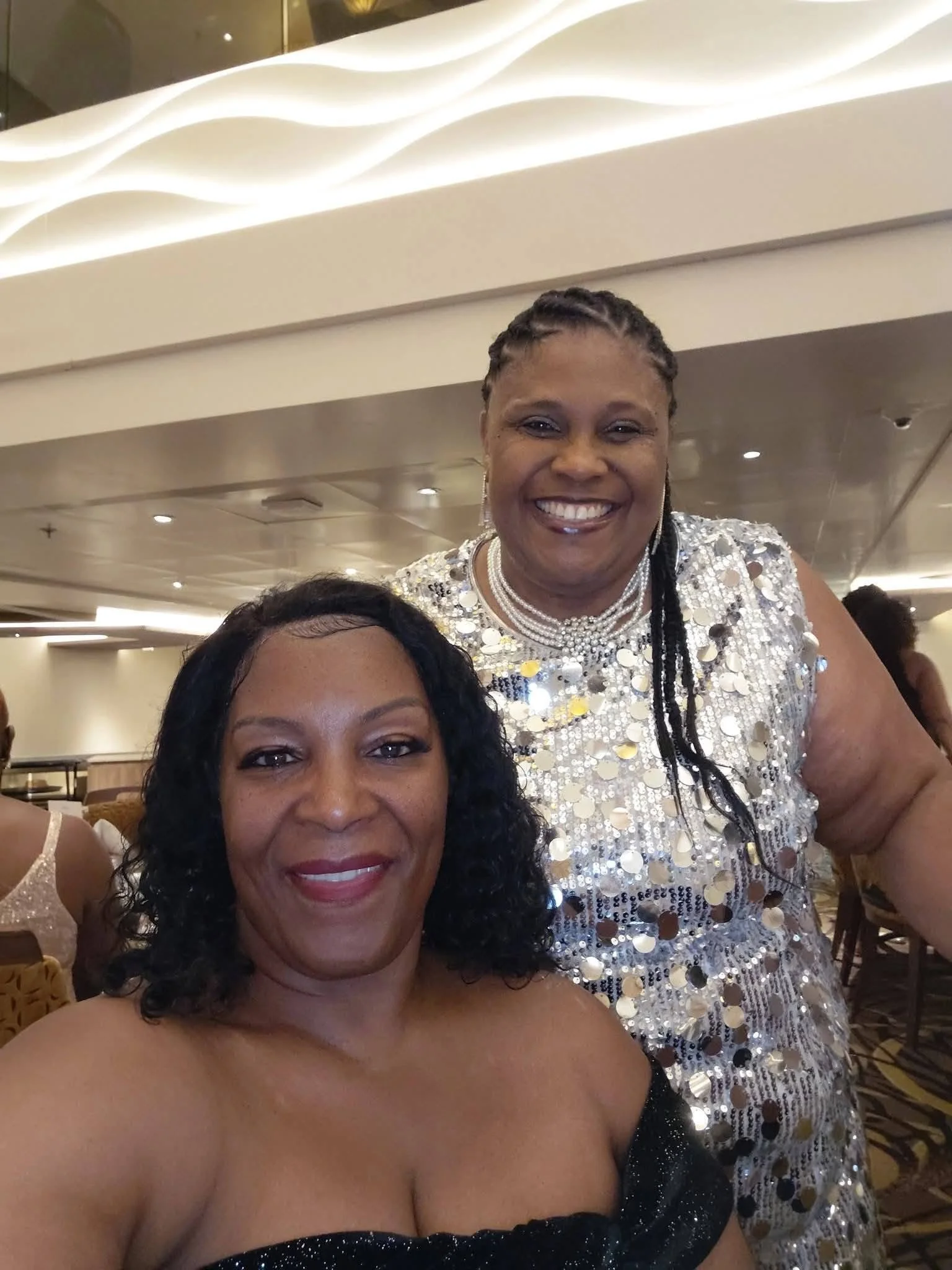 Two women smiling at an indoor event, one wearing a black strapless dress with sparkles, the other in a shiny dress with circular silver accents, and pearl necklaces, with a decorative ceiling and other attendees in the background.