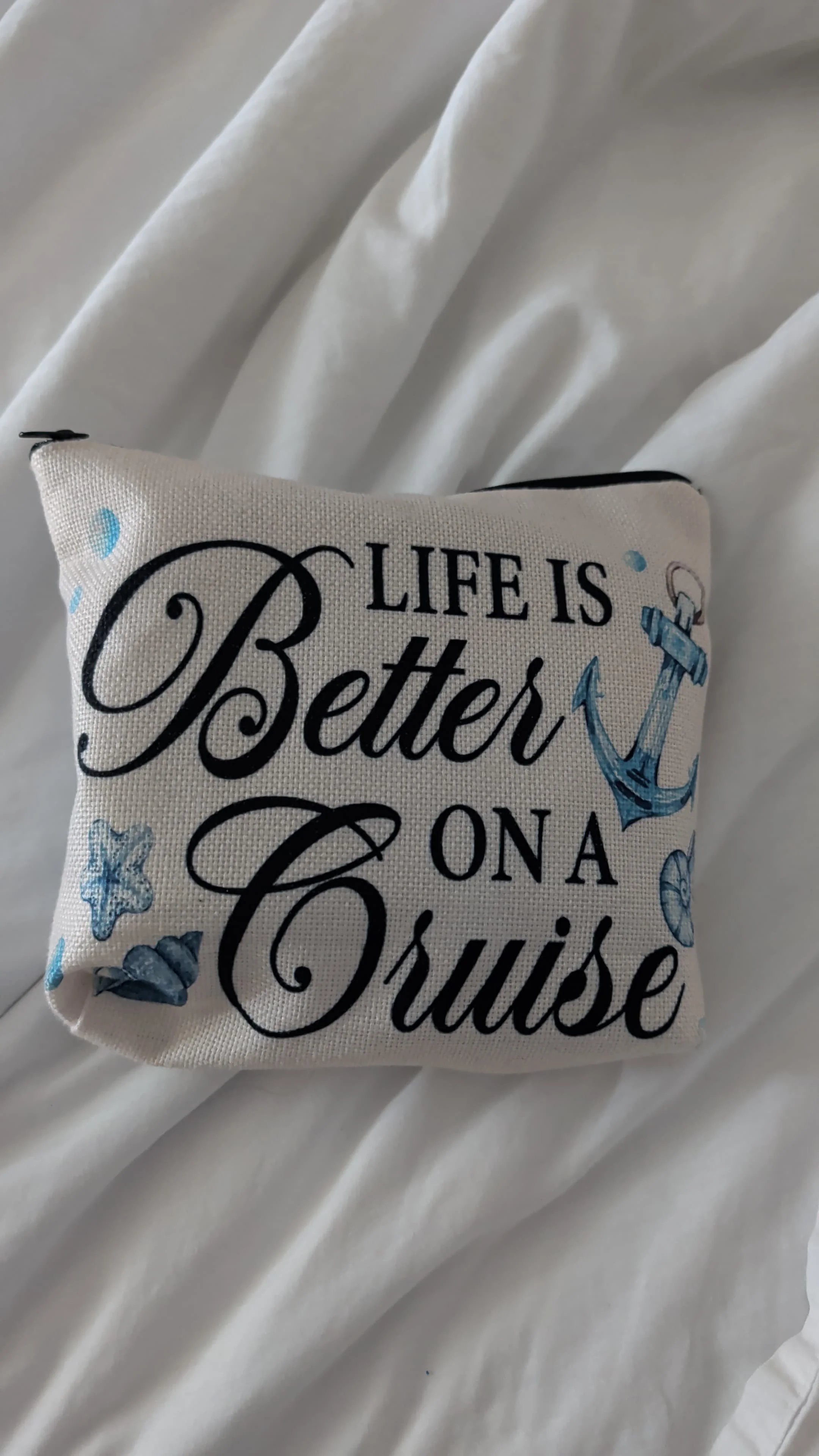 A decorative pillow with a nautical theme, featuring blue stars, shells, and nautical elements, with the phrase 'Life is Better on a Cruise' written in black cursive and block letters, placed on a white bedsheet.