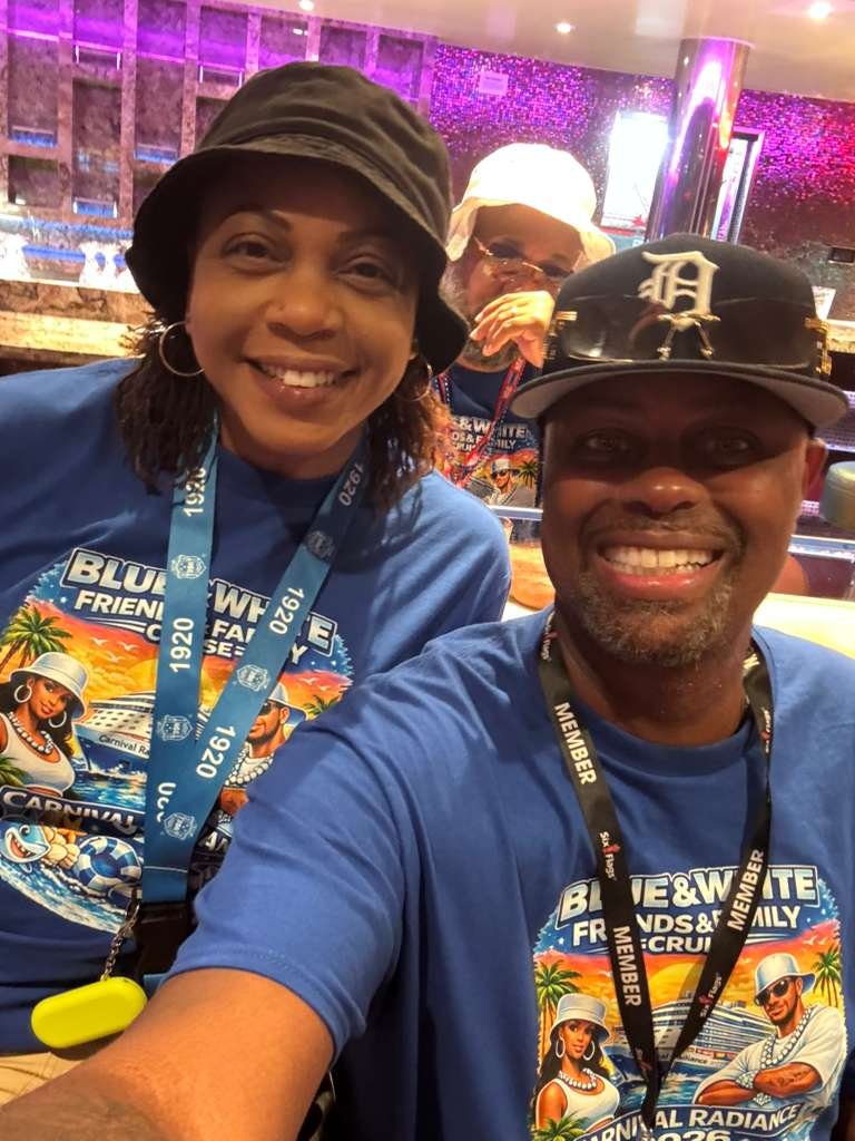 Three people taking a selfie at a cruise event, all wearing blue event T-shirts with tropical and cruise-themed graphics, and lanyards. The background shows a colorful, glittery decor.