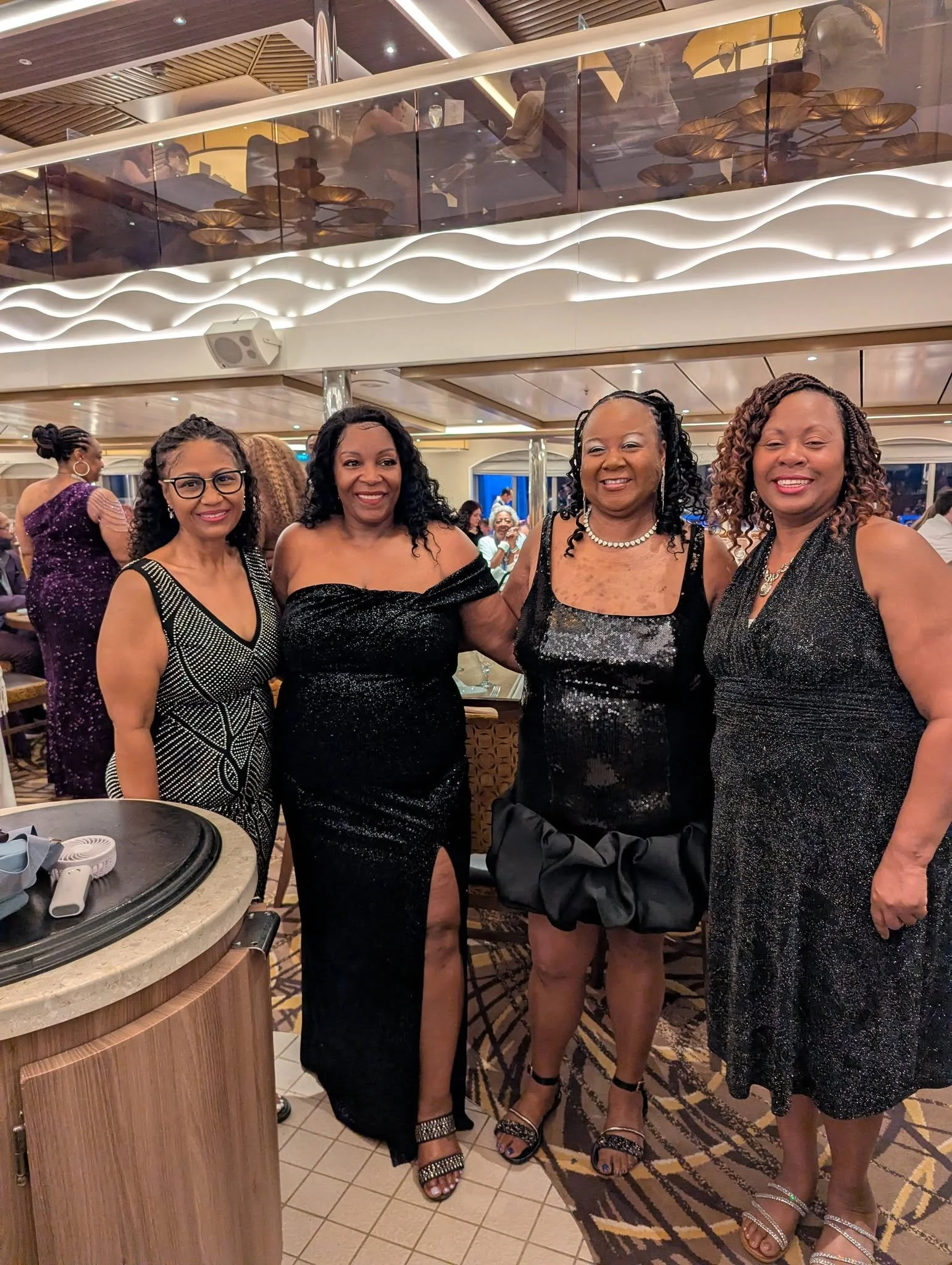 Four women dressed in black and glittery dresses posing for a group photo at a formal event in a restaurant or banquet hall. They are smiling, standing close together, with a wooden bar and a patterned carpet in the background.
