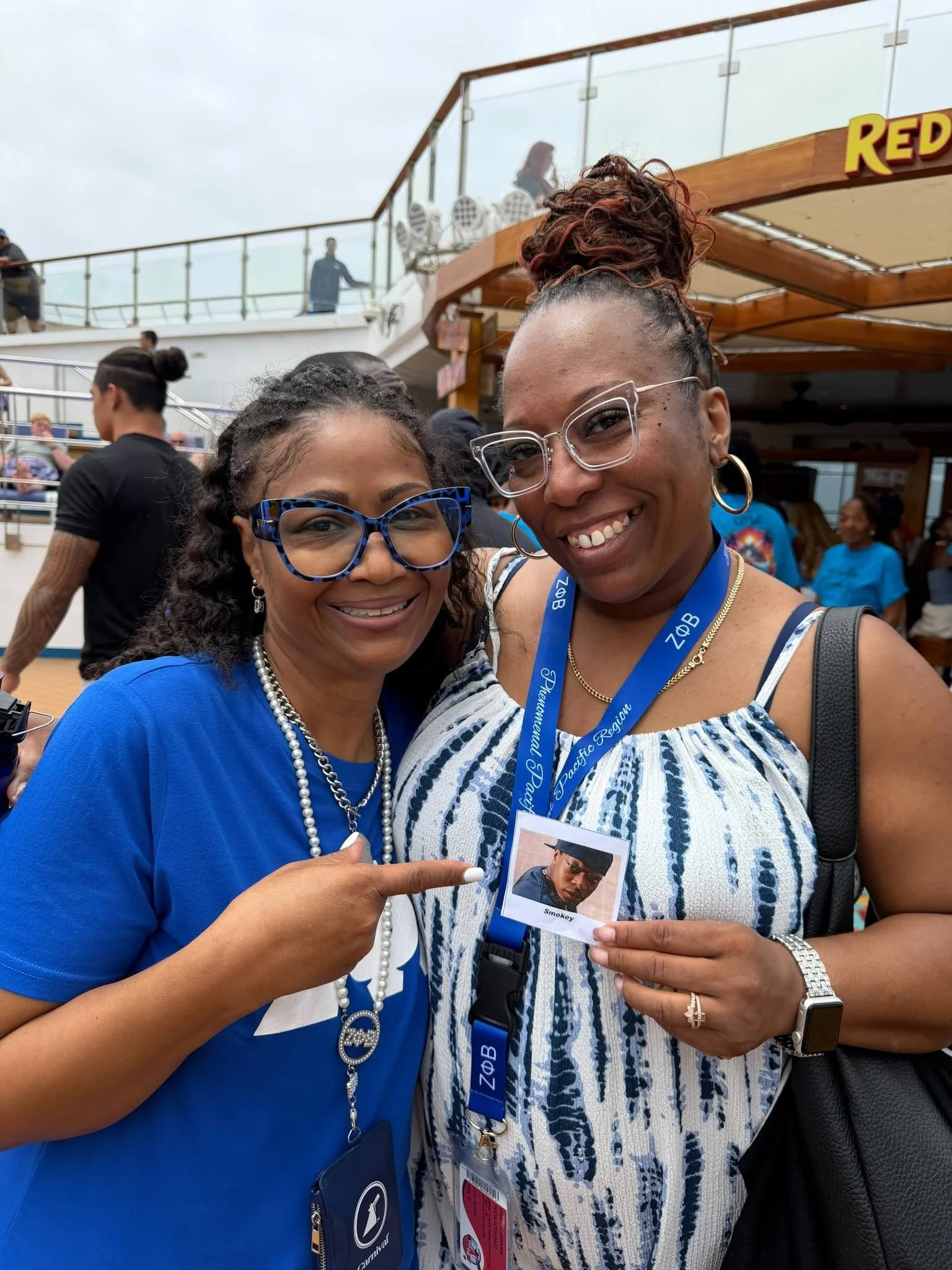 Two women smiling at the camera on a cruise ship deck, one holding a photo ID with a man's picture and the name 'Smokey', surrounded by other passengers.