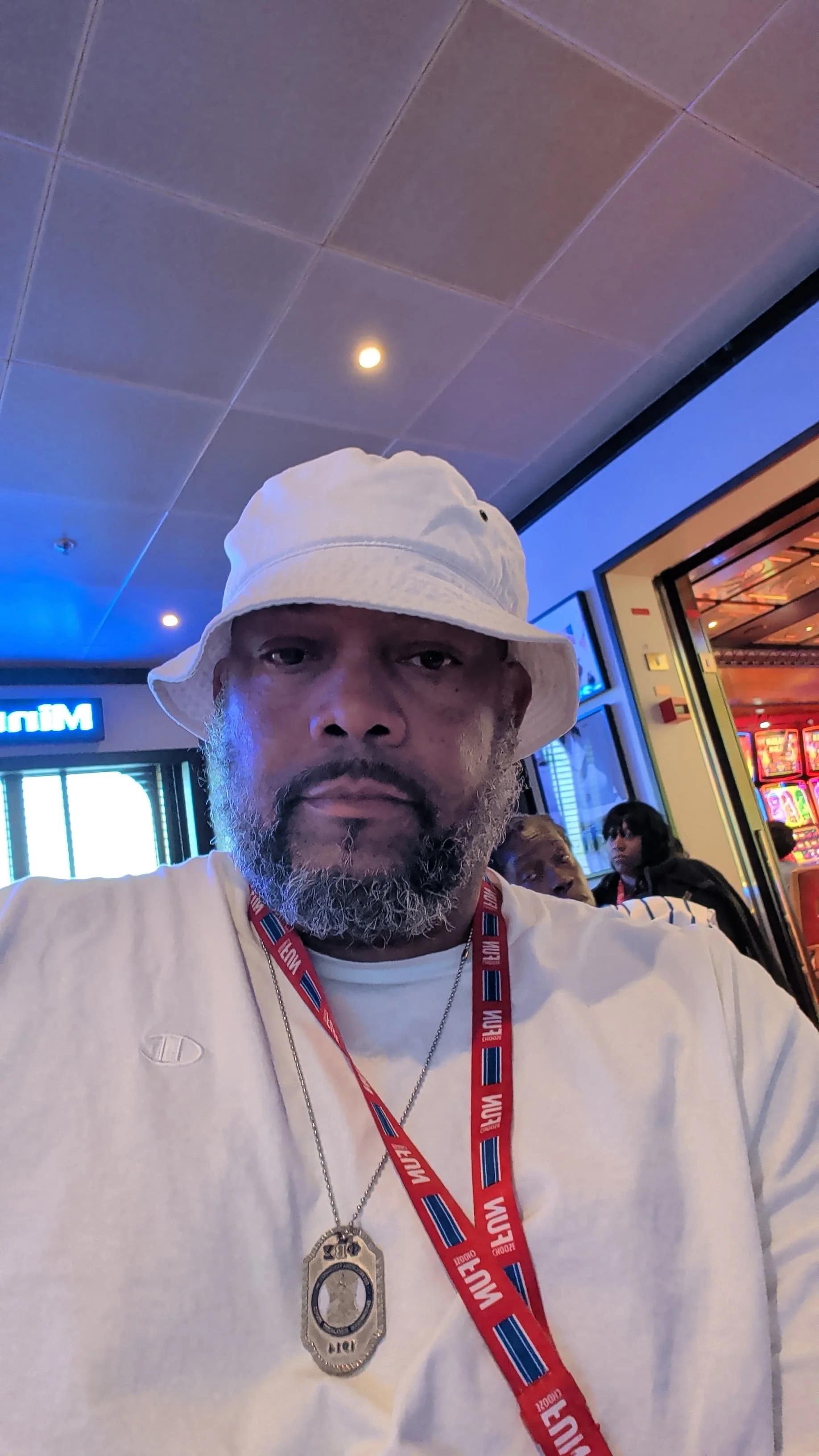 A man wearing a white bucket hat and a white T-shirt, with a badge and a red, white, and blue lanyard, sitting in a lively indoor setting with slot machines and other people in the background.