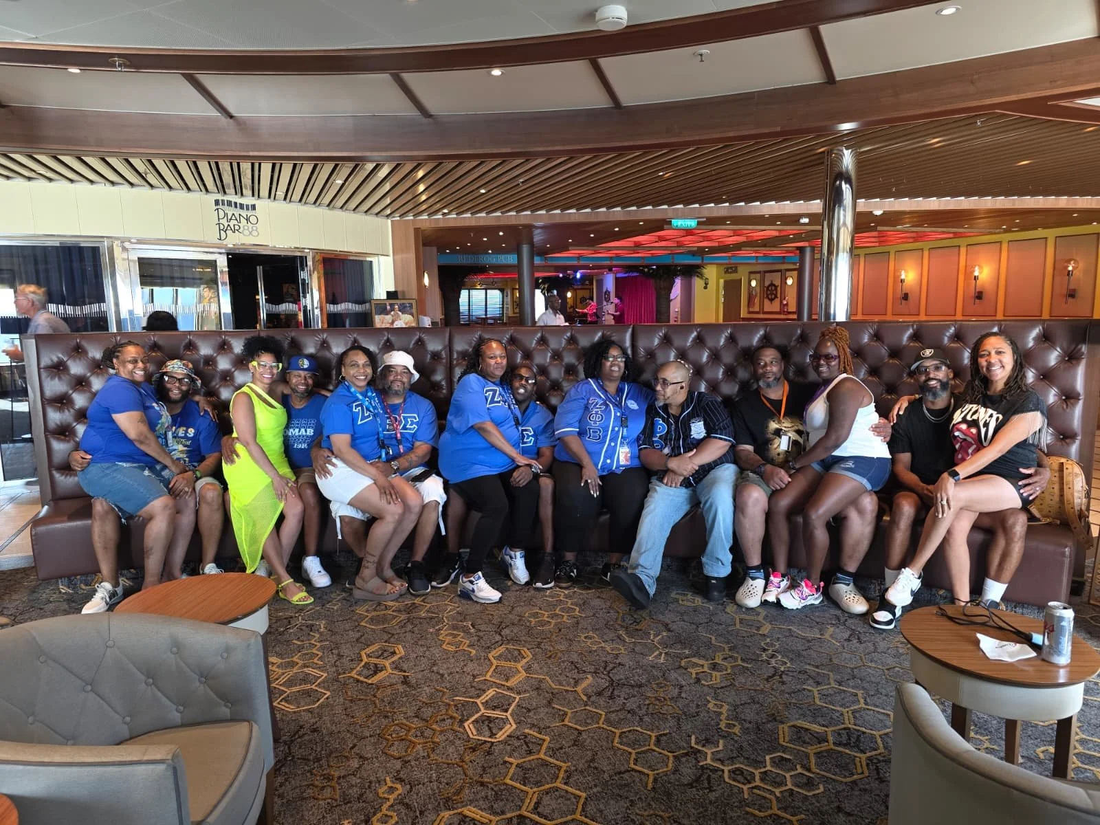 Group of people sitting on a leather couch in a restaurant or lounge area. Some individuals are wearing matching blue shirts with Greek letters. The background features wooden accents, a bar, and a stage with a performer.