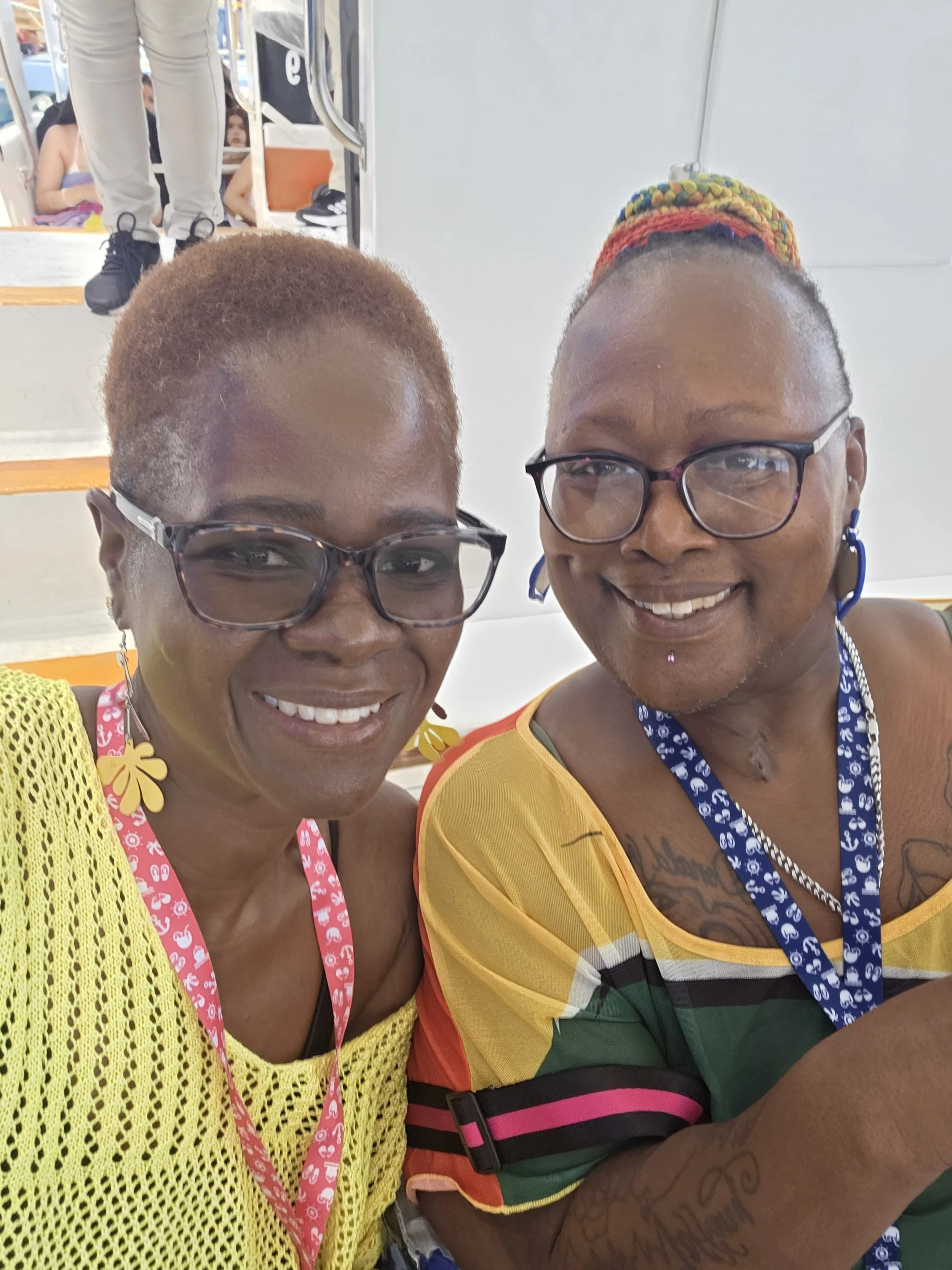 Two women taking a selfie on a train, smiling at the camera. The woman on the left has short reddish hair, glasses, and is wearing a yellow crochet top. The woman on the right has short hair with a colorful headwrap, glasses, and a yellow, green, and