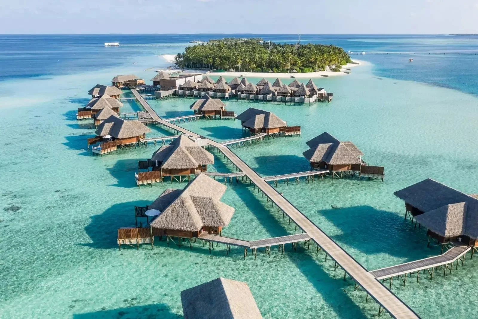 Overwater bungalows with thatched roofs connected by wooden walkways in clear turquoise water near a lush green island in a tropical setting.