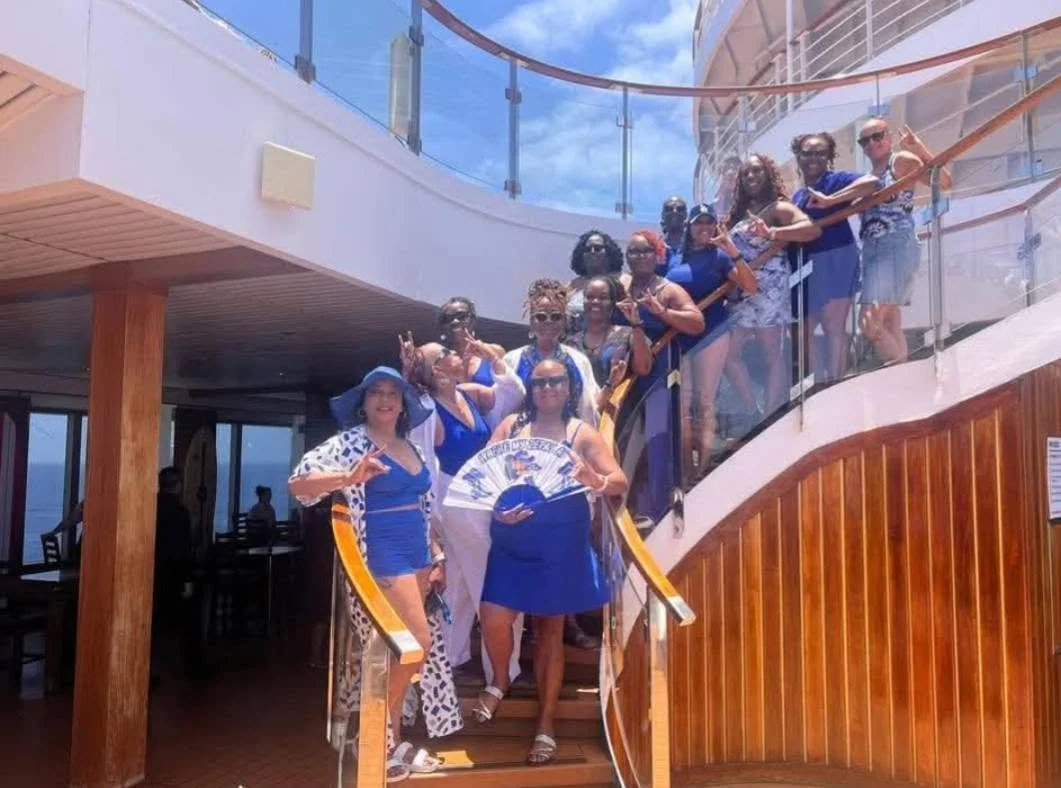 Group of women in summer dresses posing on a cruise ship staircase, with glass railings and blue sky in the background.
