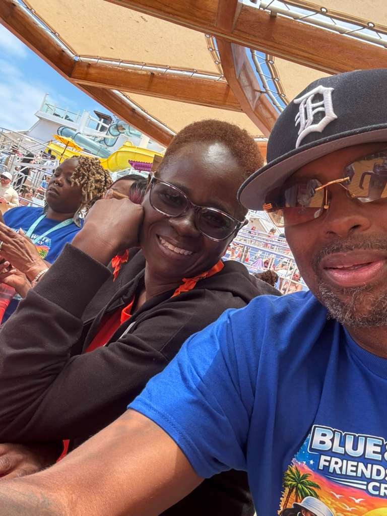 Two people smiling on a cruise ship, with amusement park rides and other passengers in the background.