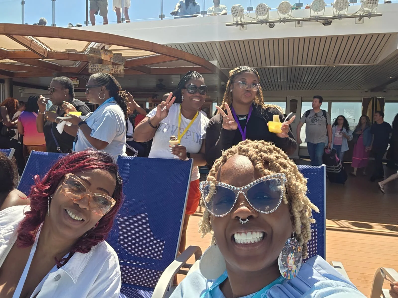 Smiling women taking a selfie on a cruise ship's deck with other passengers in the background.