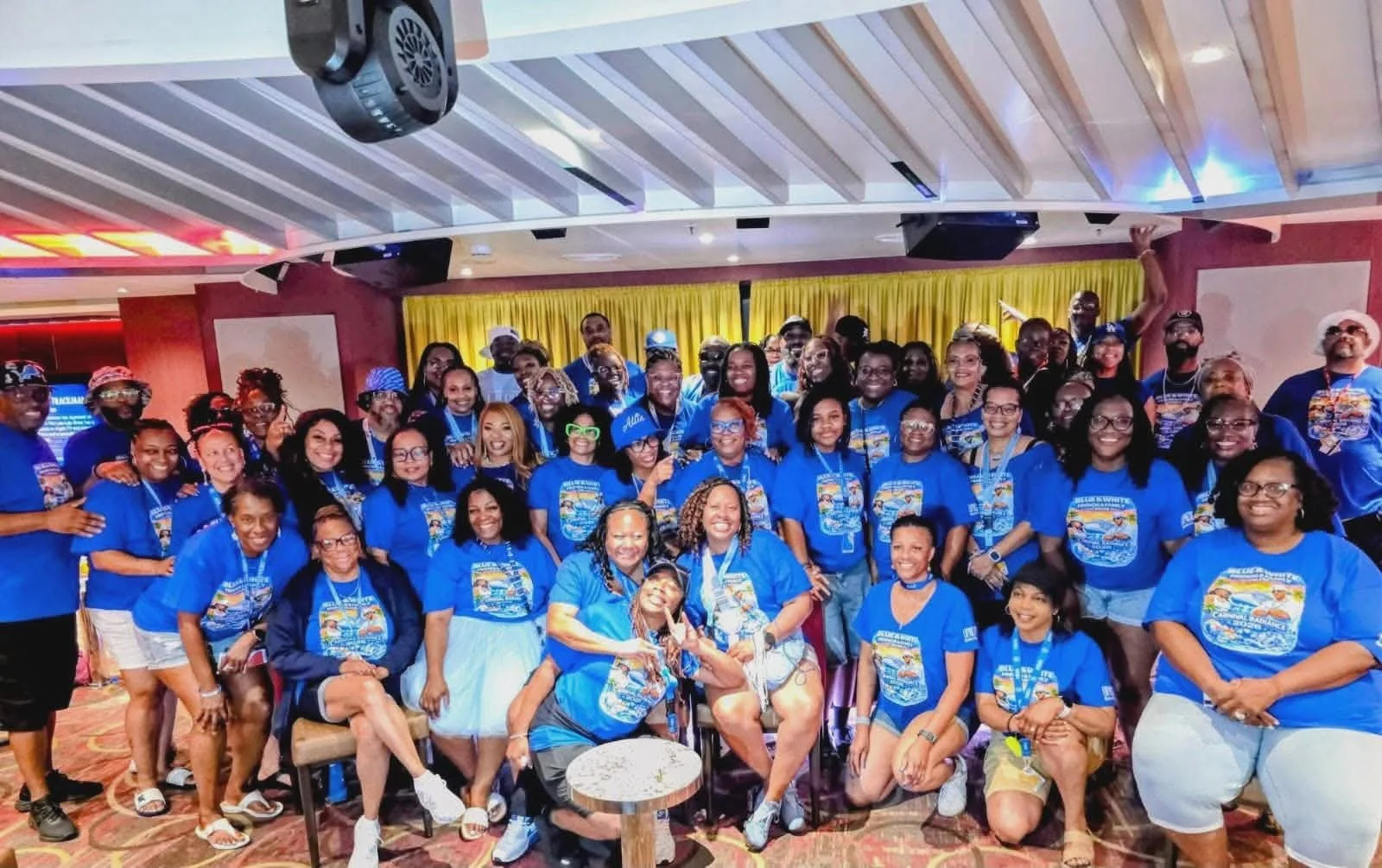 A large group of people gathered indoors for a group photo, all wearing matching blue T-shirts with a graphic design on the front, with some seated and others standing. The background features yellow curtains and a ceiling with white panels.