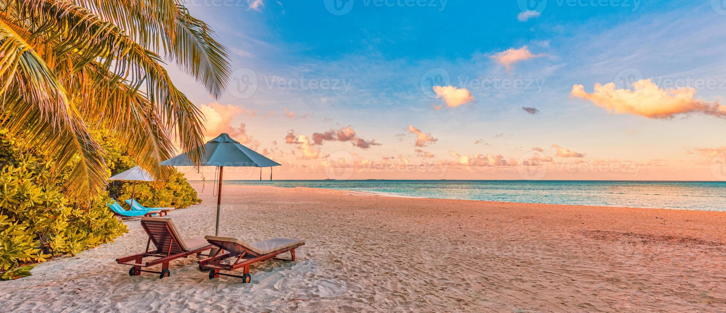 Beach scene at sunset with palm trees, beach chairs under umbrellas, sandy shore, and calm ocean with pink clouds in the sky.