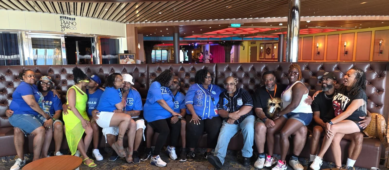 Group of people sitting on a tufted leather booth in a restaurant or lounge, some wearing casual clothing, some in matching blue shirts, with a tropical or cruise ship-themed decor in the background.