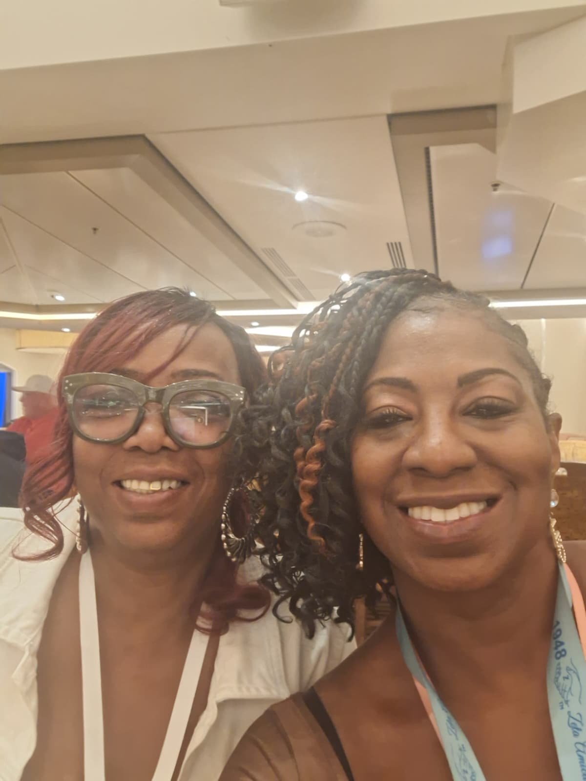Two women smiling and taking a selfie in an indoor setting with ceiling lights and ventilation system visible.