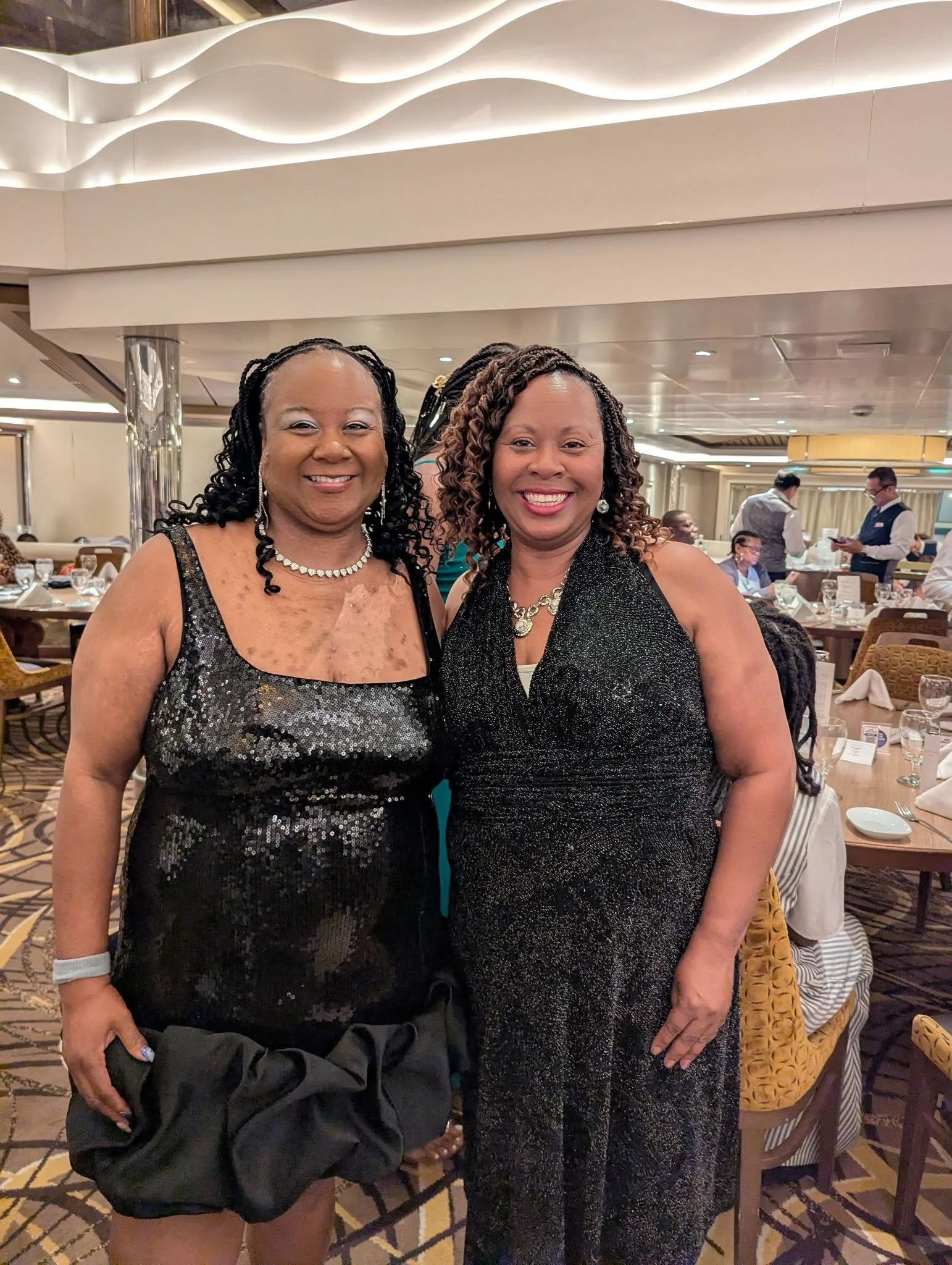 Two women in elegant black dresses posing together at a banquet hall, smiling for the camera. Other guests and servers are visible in the background.