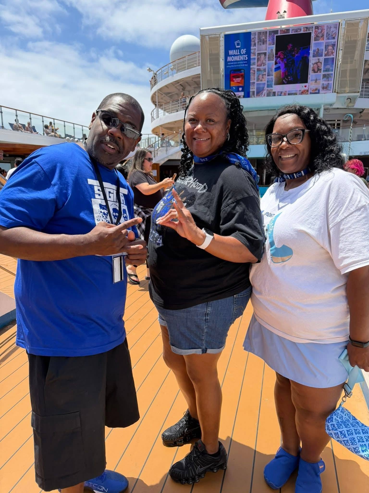 Three people posing on the deck of a cruise ship, with a large digital screen in the background displaying photos and the text "Wall of Moments." The man on the left is wearing a blue shirt, sunglasses, and pointing with his right hand. The woman in 