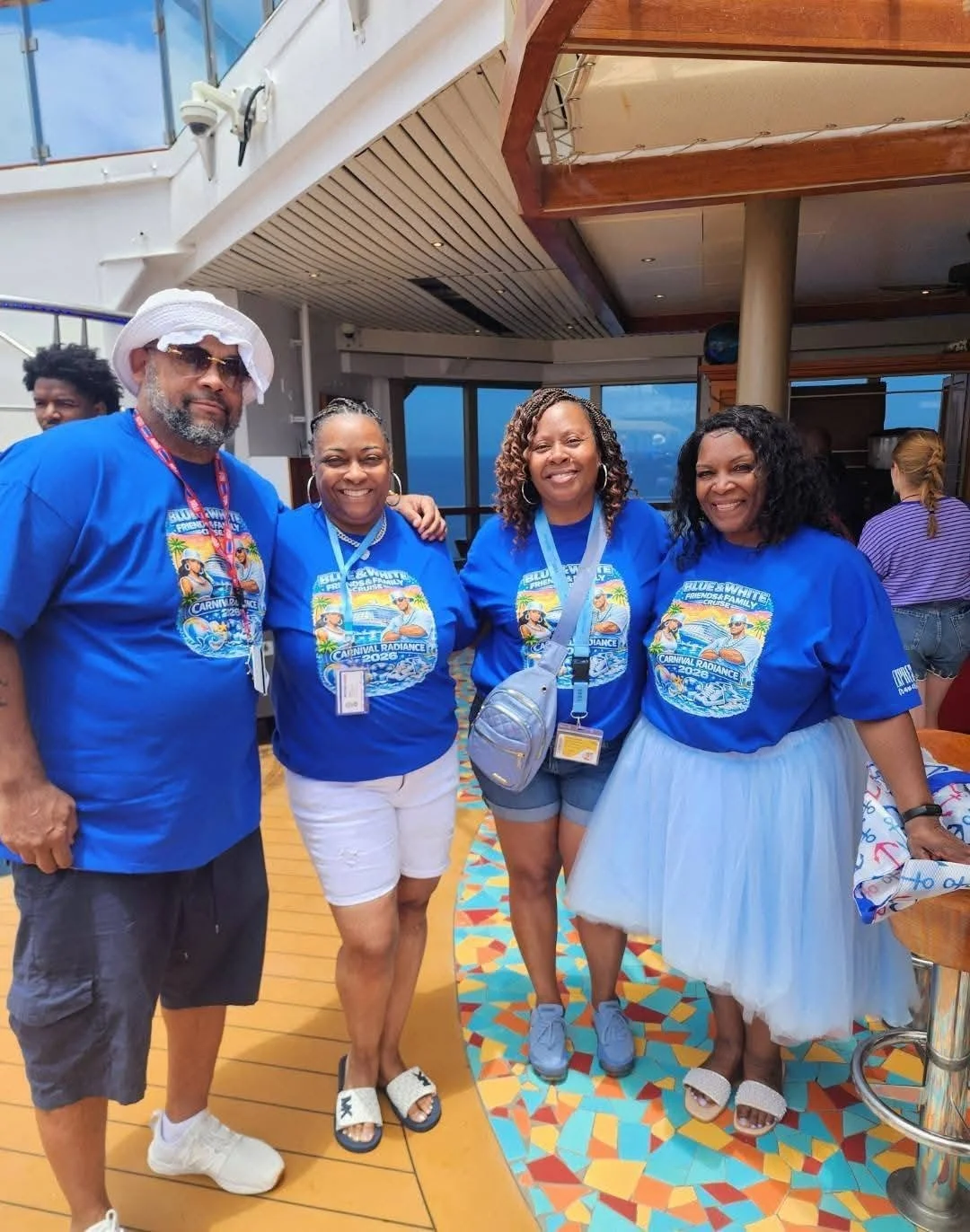 Four people on a cruise ship deck wearing matching blue T-shirts with a tropical cruise design and event information, smiling for the photo.