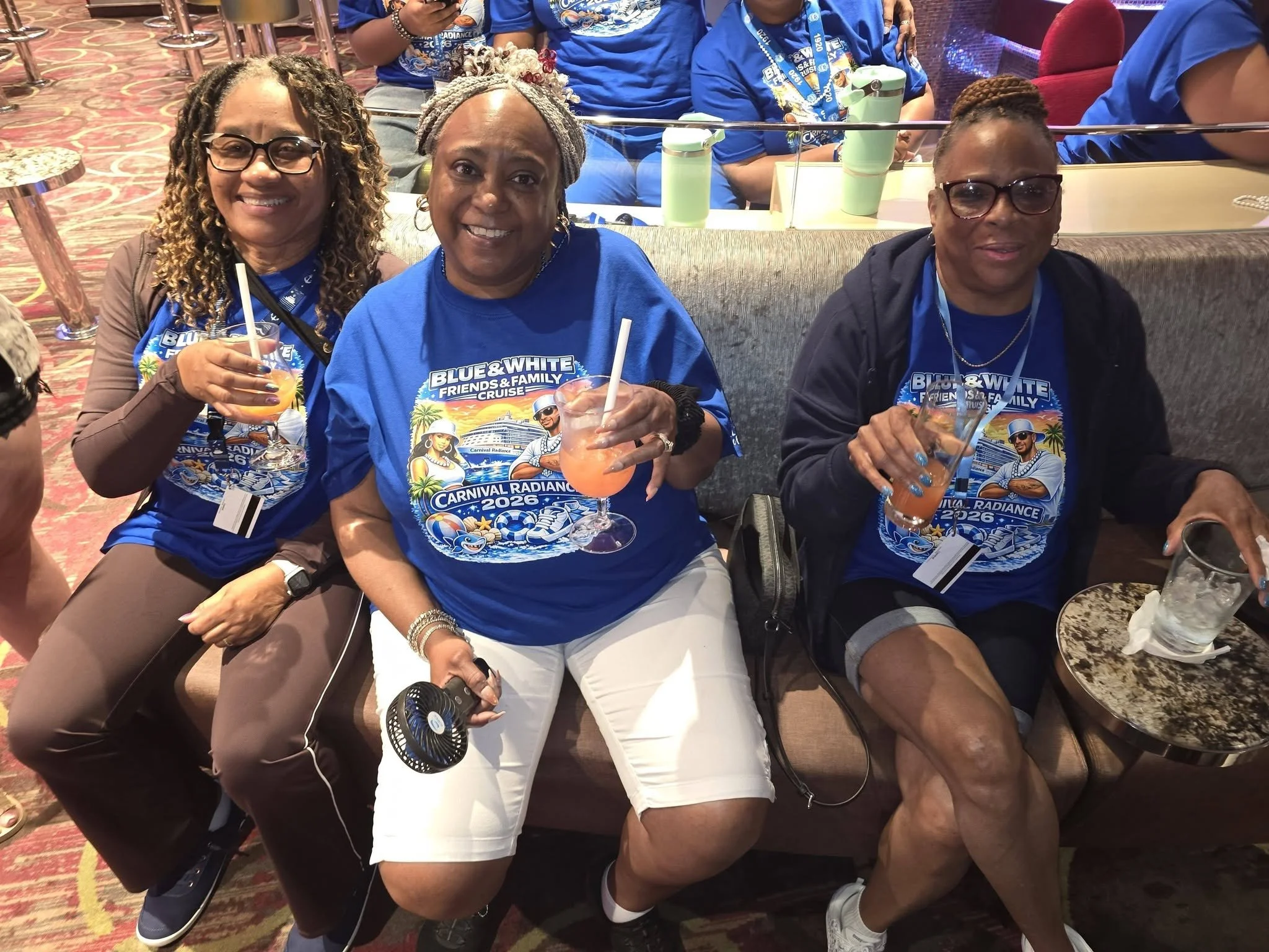 Three women sitting together, smiling, at a social event or cruise, wearing blue shirts that say "Blue & White Friends & Family Cruise 2026" and holding drinks. They are indoors with a decorative carpet and other attendees visible in the background.