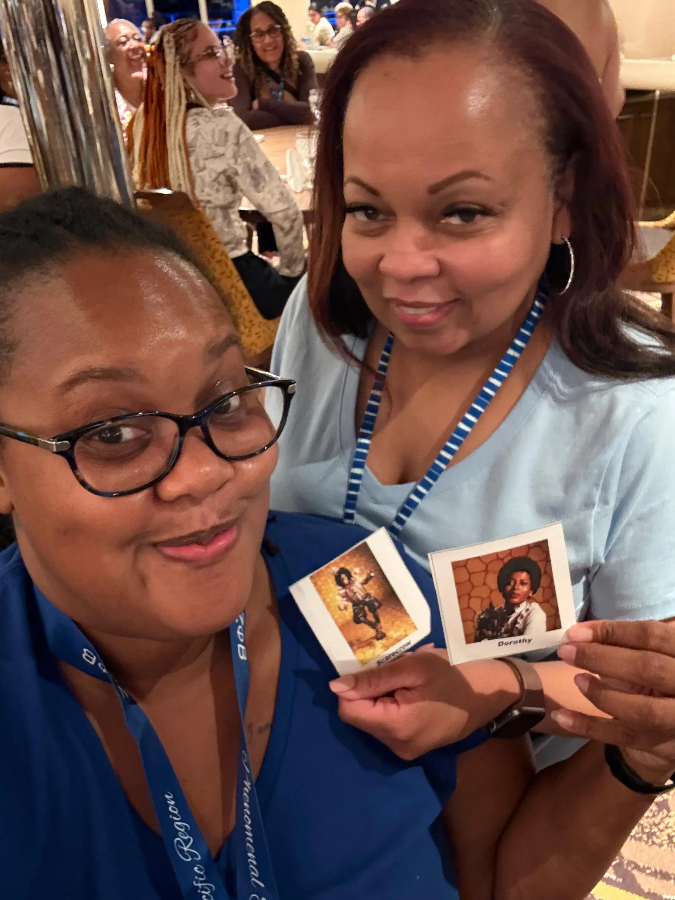 Two women taking a selfie at a social event, holding cards with images of a woman in a Western outfit and the name 'Dorothy'. In the background, other people are seated, talking, and laughing.