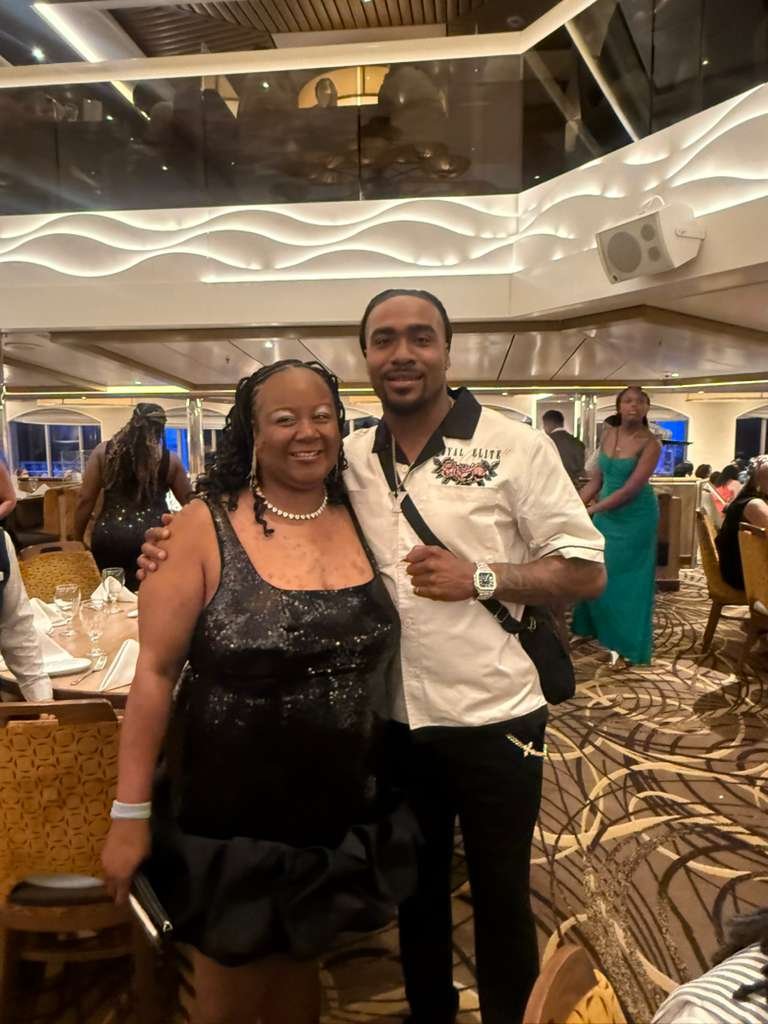 A woman and a man smiling and posing together at a formal event in a banquet hall with decorative lighting and people seated at tables.