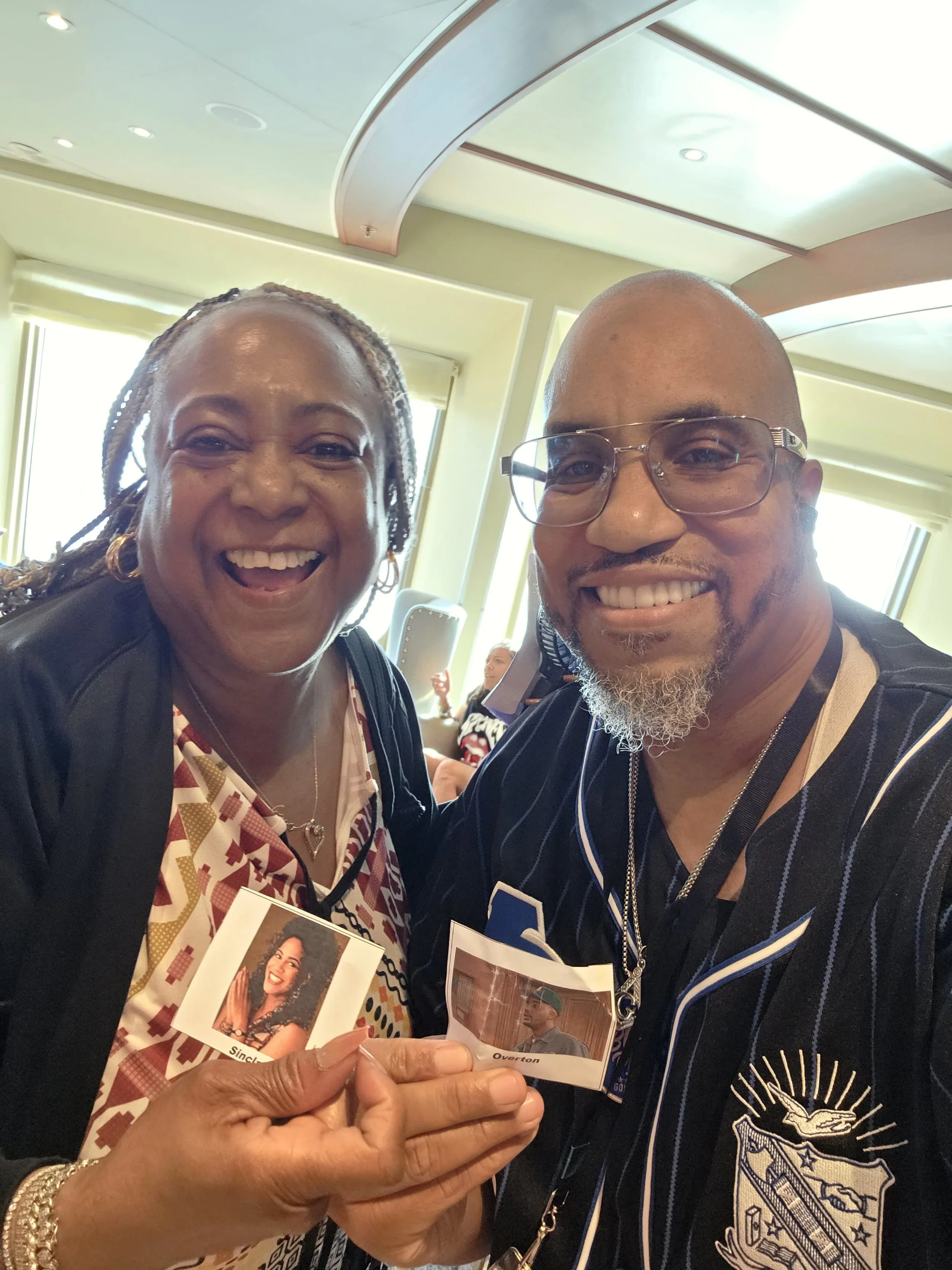 Smiling African American woman and man taking a selfie inside a well-lit conference room, holding photographs.