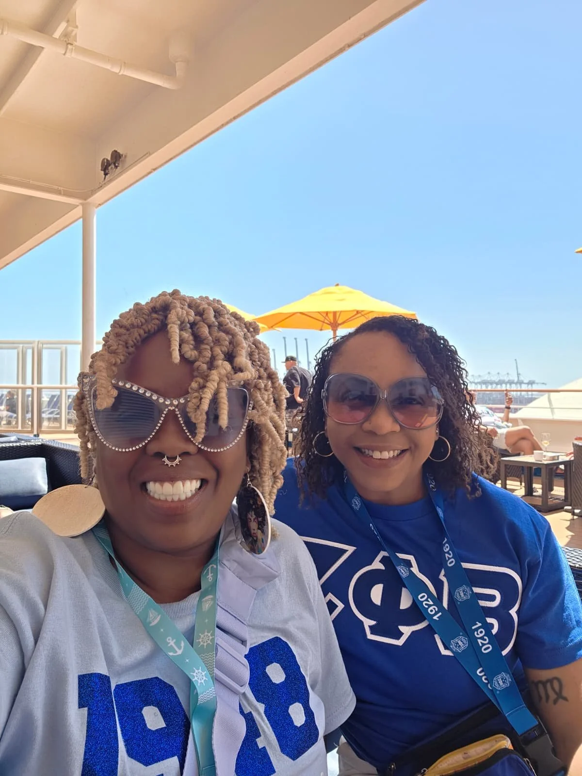 Two women smiling at a sunny outdoor venue, wearing large sunglasses, blue and white sports jerseys, and event lanyards. One woman has blonde dreadlocks, and the other has curly hair.