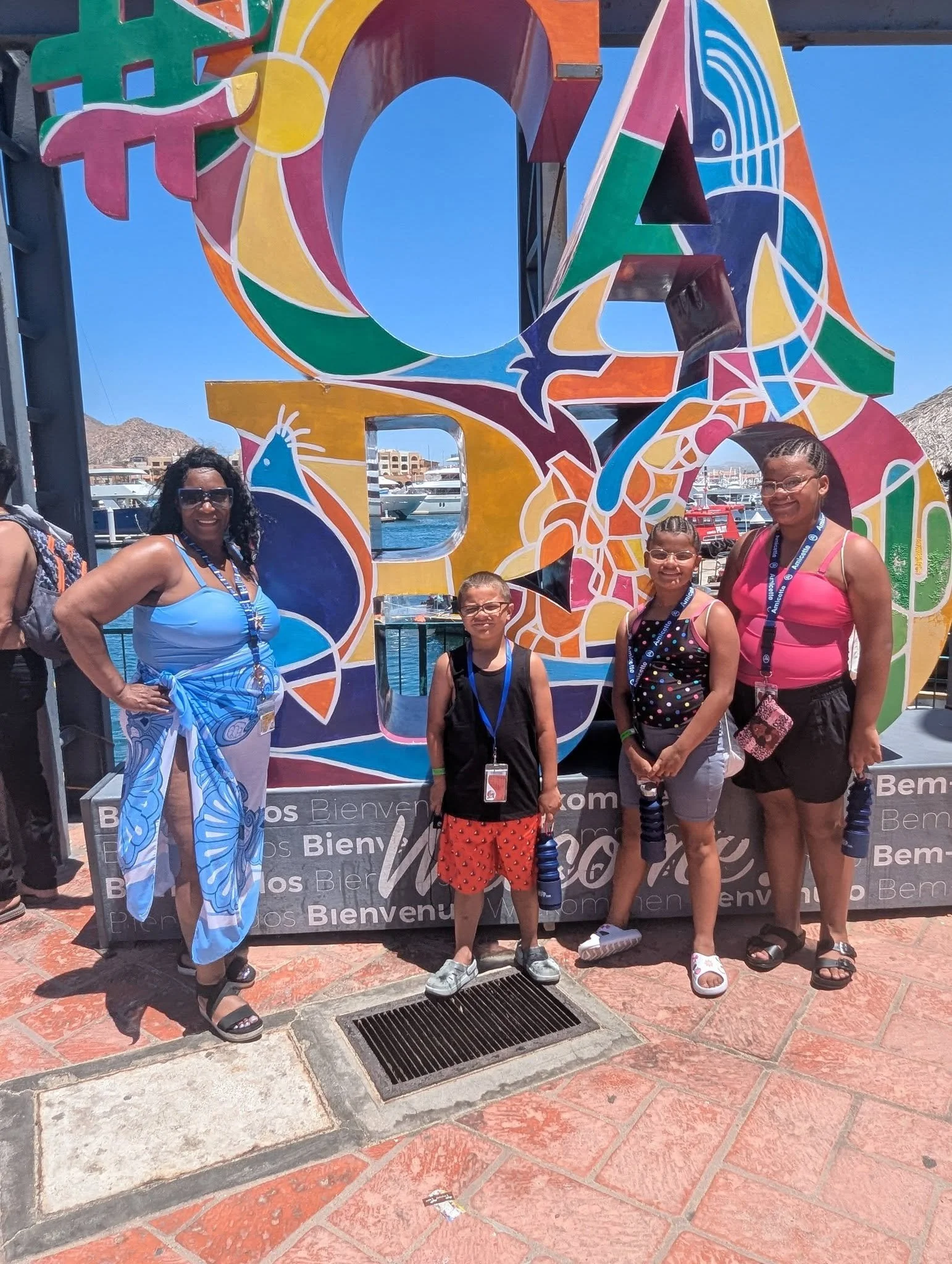 A group of five people, consisting of two adult women and three children, posing in front of a colorful sign that says ''Bienvenido'' on a sunny day at a marina or harbor with boats docked in the background.