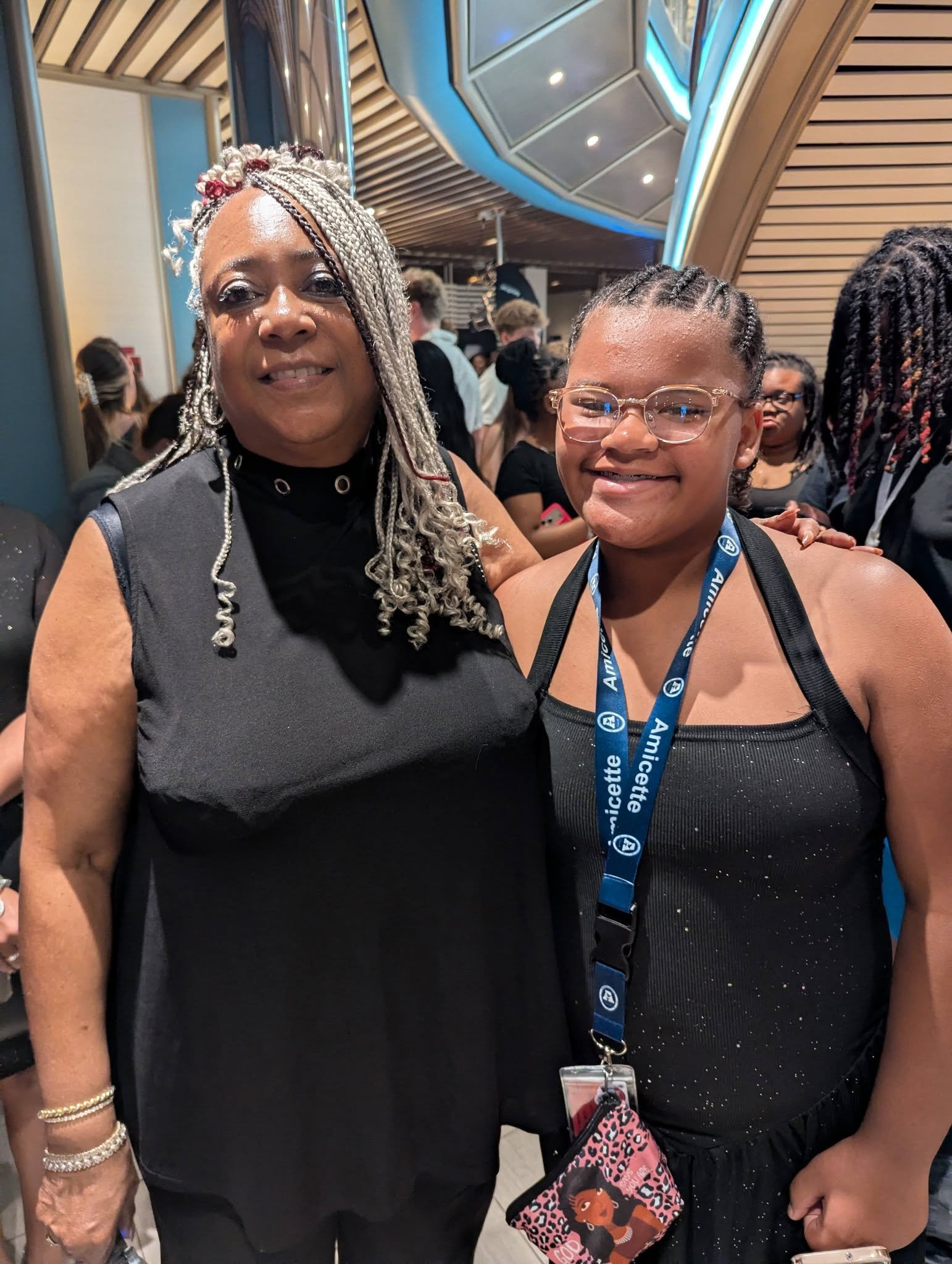 A woman with long, braided silver hair and a woman with short, braided hair and glasses pose together at an indoor event. They are surrounded by other people and the background features wooden and metallic architectural elements with blue accent ligh