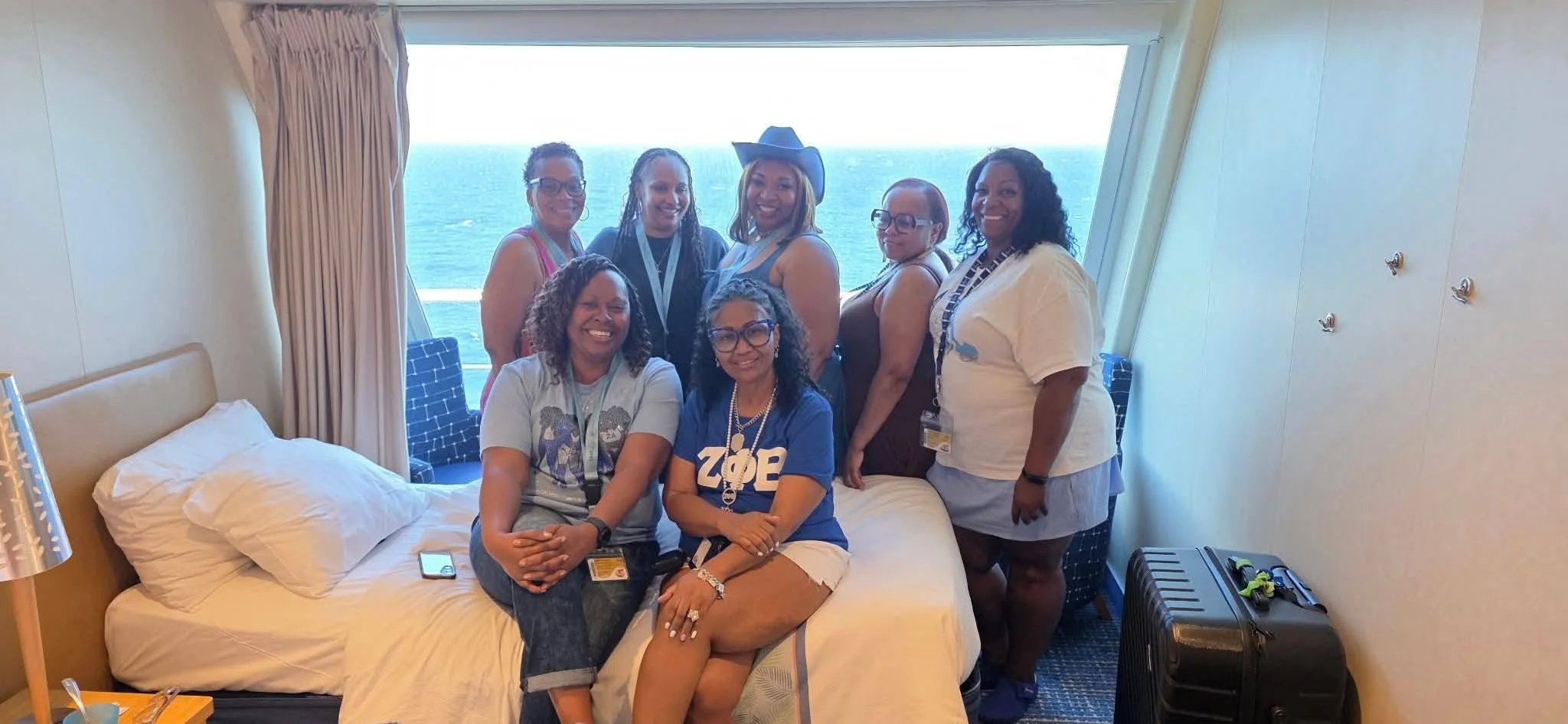 Group of women on a cruise ship deck with ocean in the background, some wearing name tags and one with a cowboy hat, smiling and posing for the photo.
