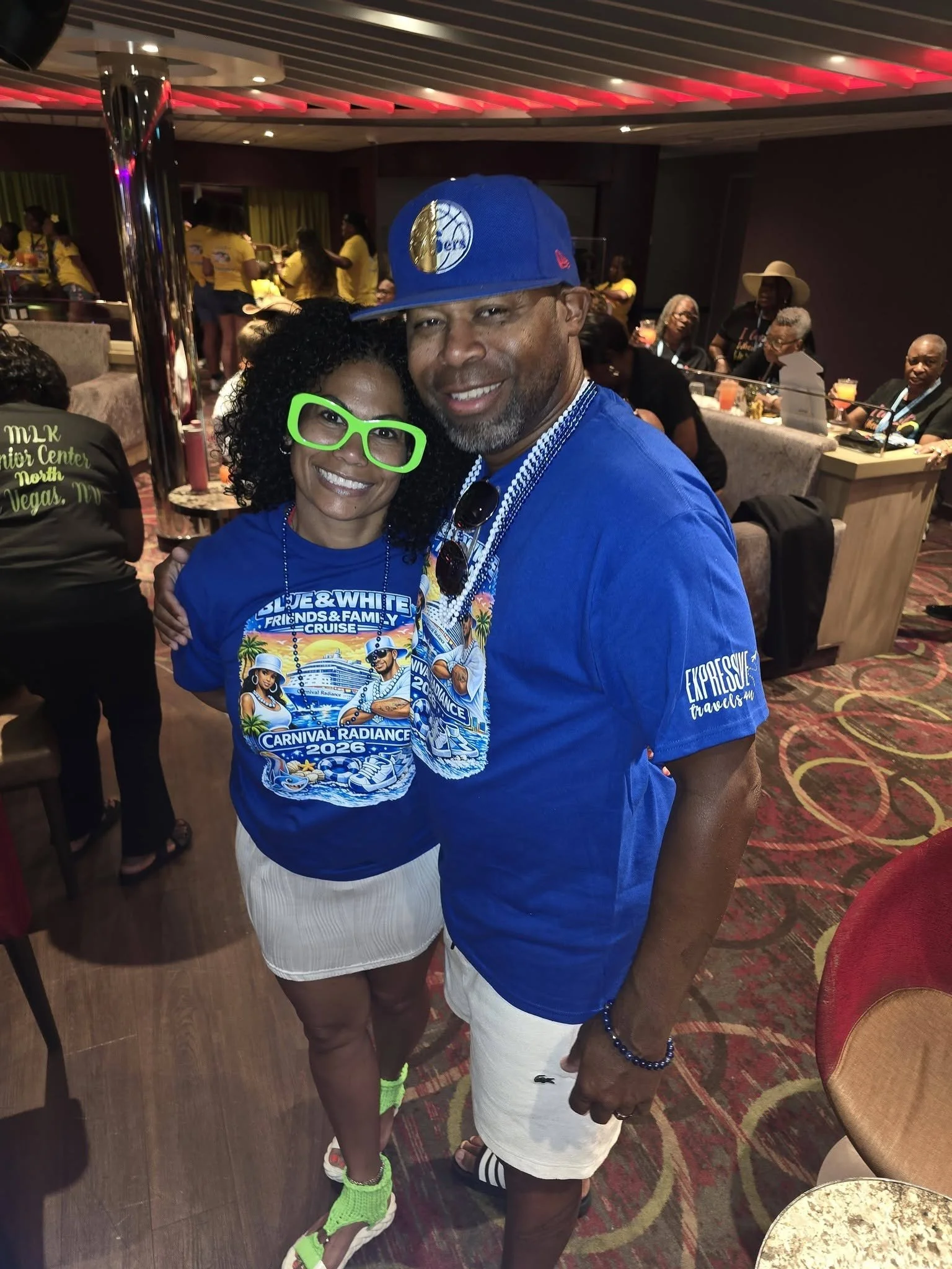 A man and woman posing together at a lively indoorEvent, with the woman wearing bright green glasses, a carnival-themed shirt, and neon green sneakers, and the man wearing a blue cap and white shorts. The background features people sitting at a bar a