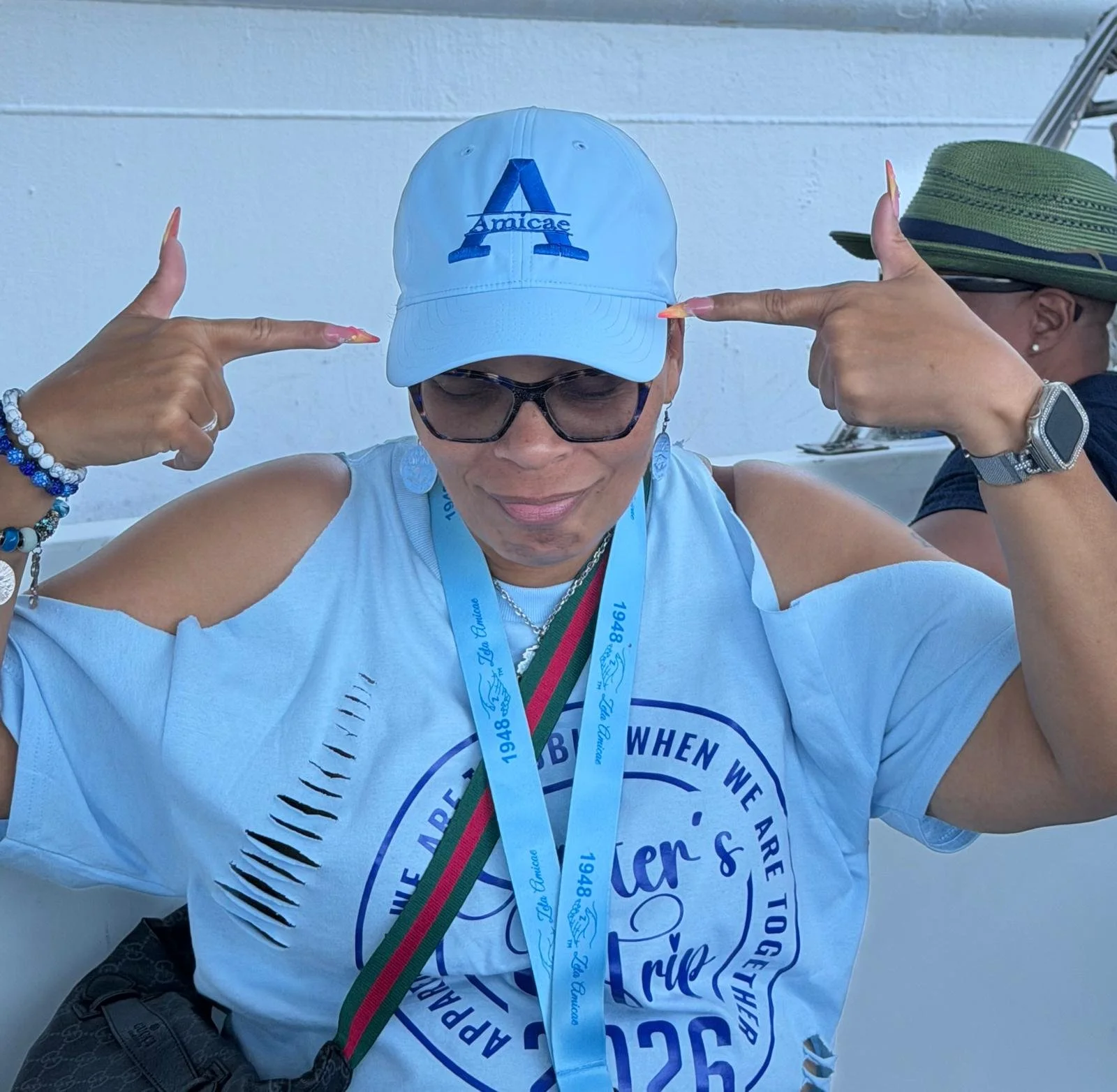 A woman wearing sunglasses, a white baseball cap with a blue 'A' and 'Amicae' embroidered, a blue and white t-shirt, and multiple medals around her neck, is pointing to her cap with both hands, in front of a white wall.