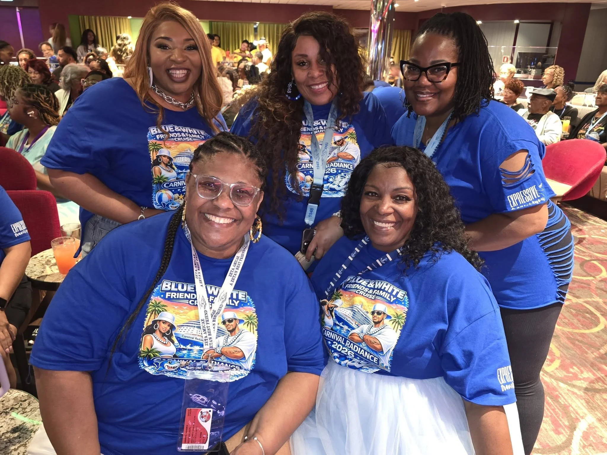 Group of women wearing matching blue t-shirts with a beach and cruise-themed design, smiling at a social event or gathering on a cruise ship or conference hall.