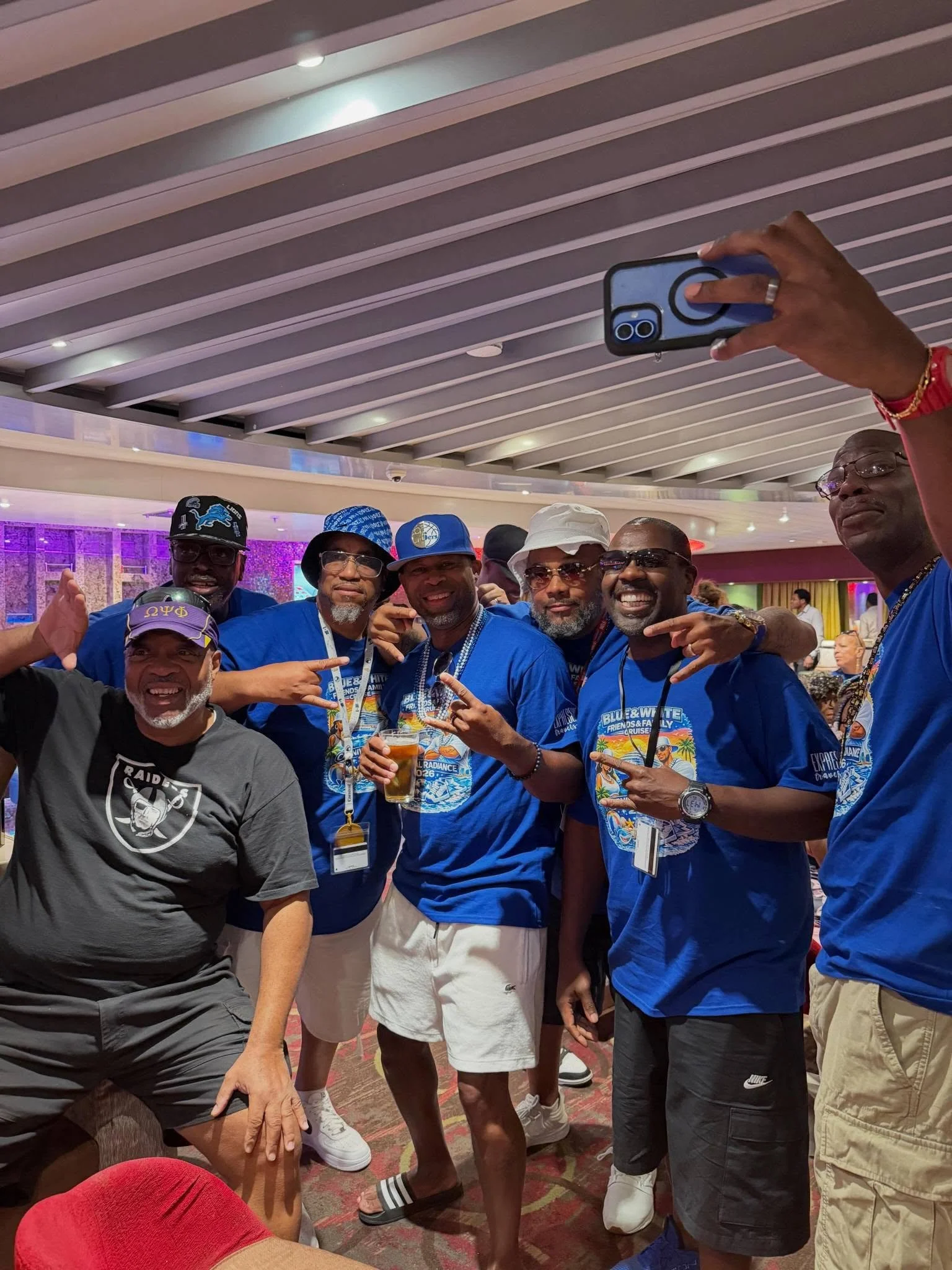 Group of men posing for a photo at an event, some making hand gestures, wearing blue shirts, hats, and sunglasses, with a colorful patterned carpet and ceiling with horizontal beams in the background.