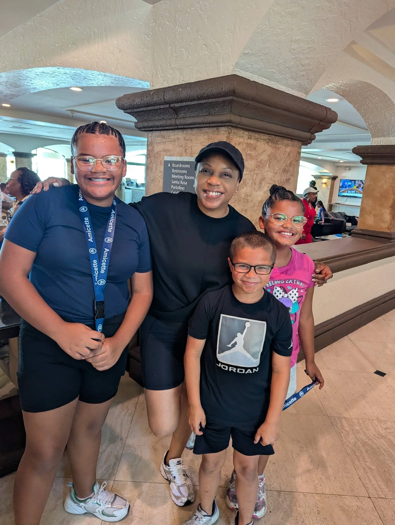 A group of children and an adult smiling at the camera inside a building. The children are wearing casual clothes and sneakers, and some are wearing glasses. The background shows a reception area with a sign listing rooms and restrooms.