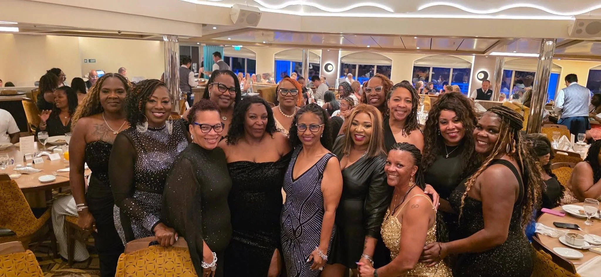 Group of women posing together at a formal event in a banquet hall with tables and other guests in the background.