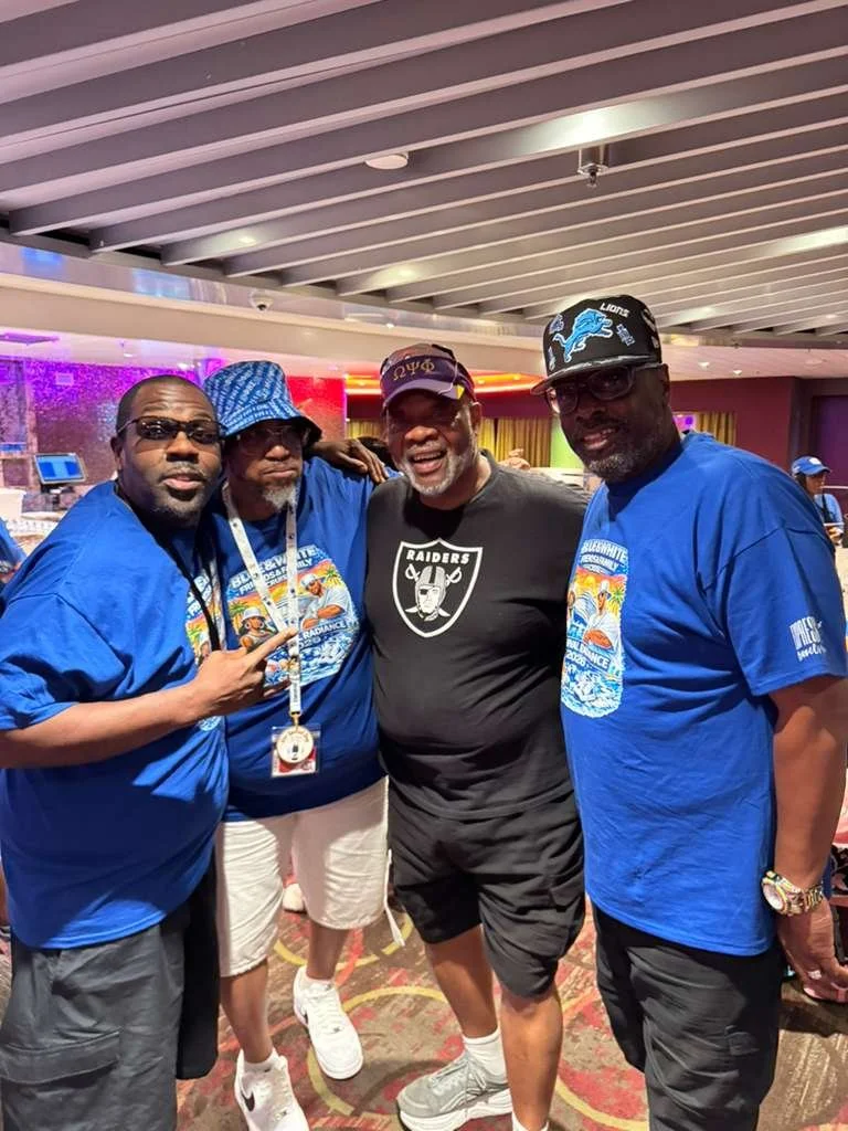 Four men posing together indoors, three wearing blue shirts and one wearing a Raiders t-shirt, with colorful lighting in the background.