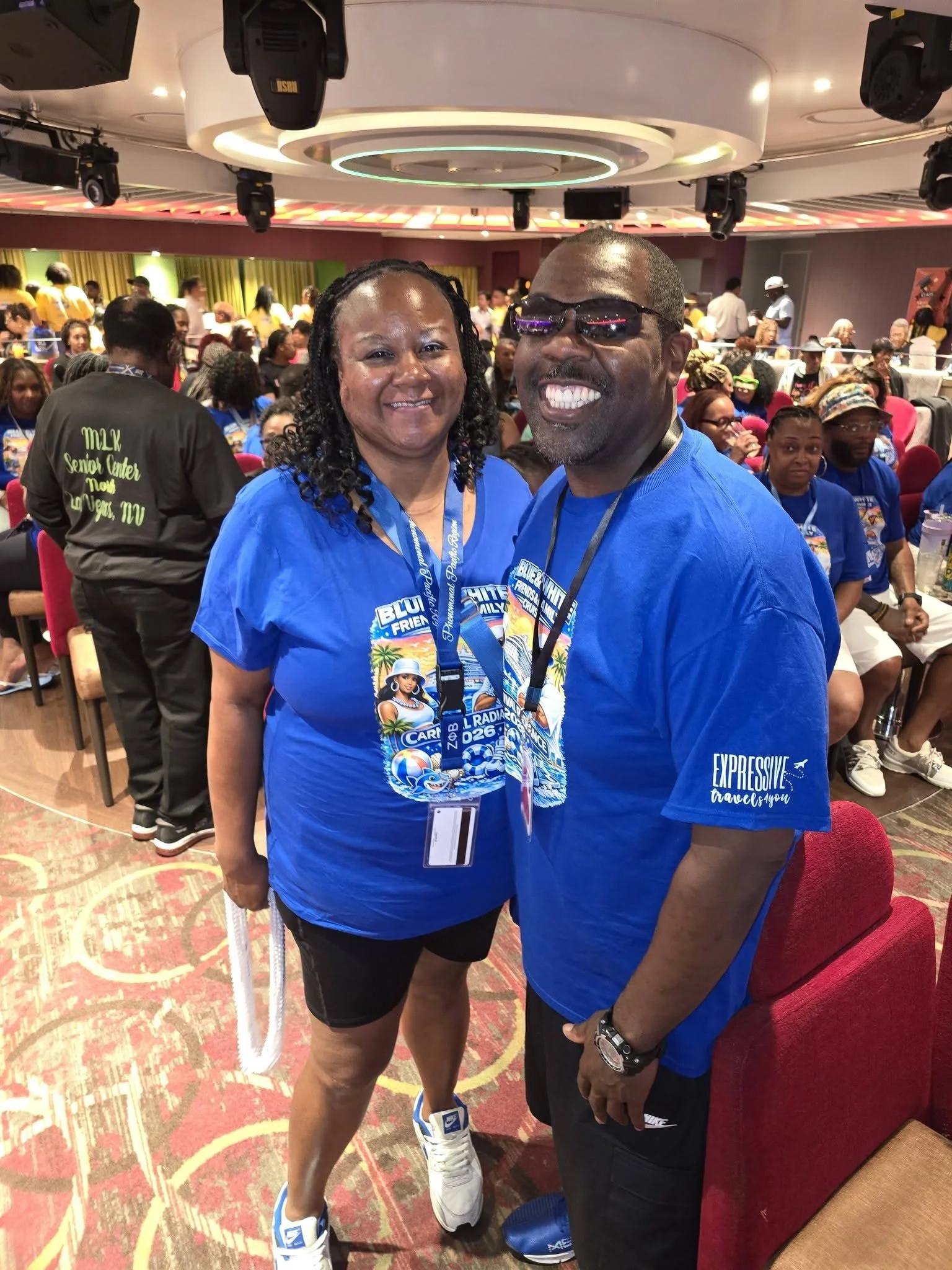 Two people smiling and posing for a photo at a crowded event. The woman on the left is wearing a blue event T-shirt, black shorts, and sneakers. The man on the right is wearing sunglasses, a blue T-shirt with white text, and black pants. The backgrou