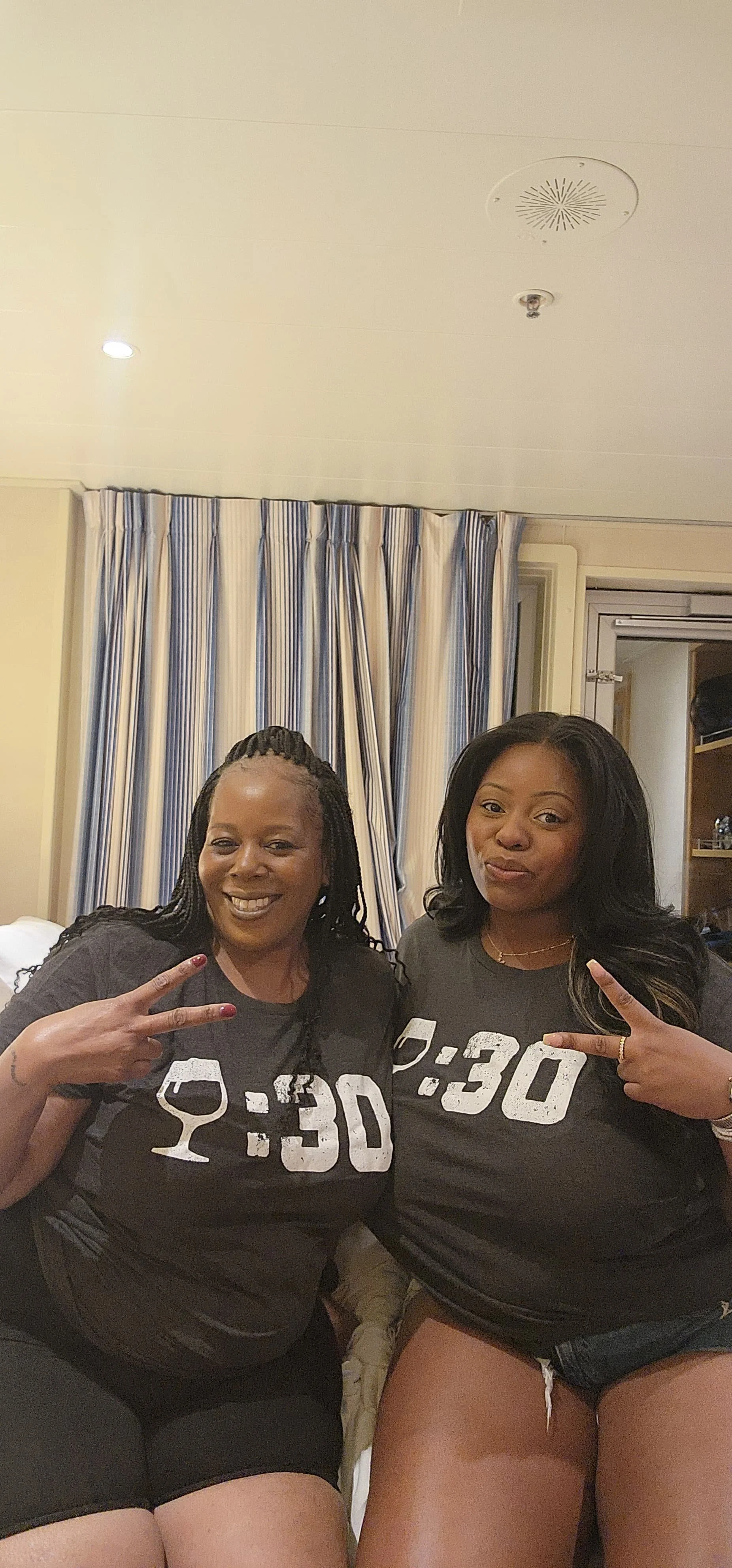 Two women wearing matching black shirts with '8:30' and a wine glass graphic, making peace signs and smiling in a hotel room with curtains and a closet in the background.