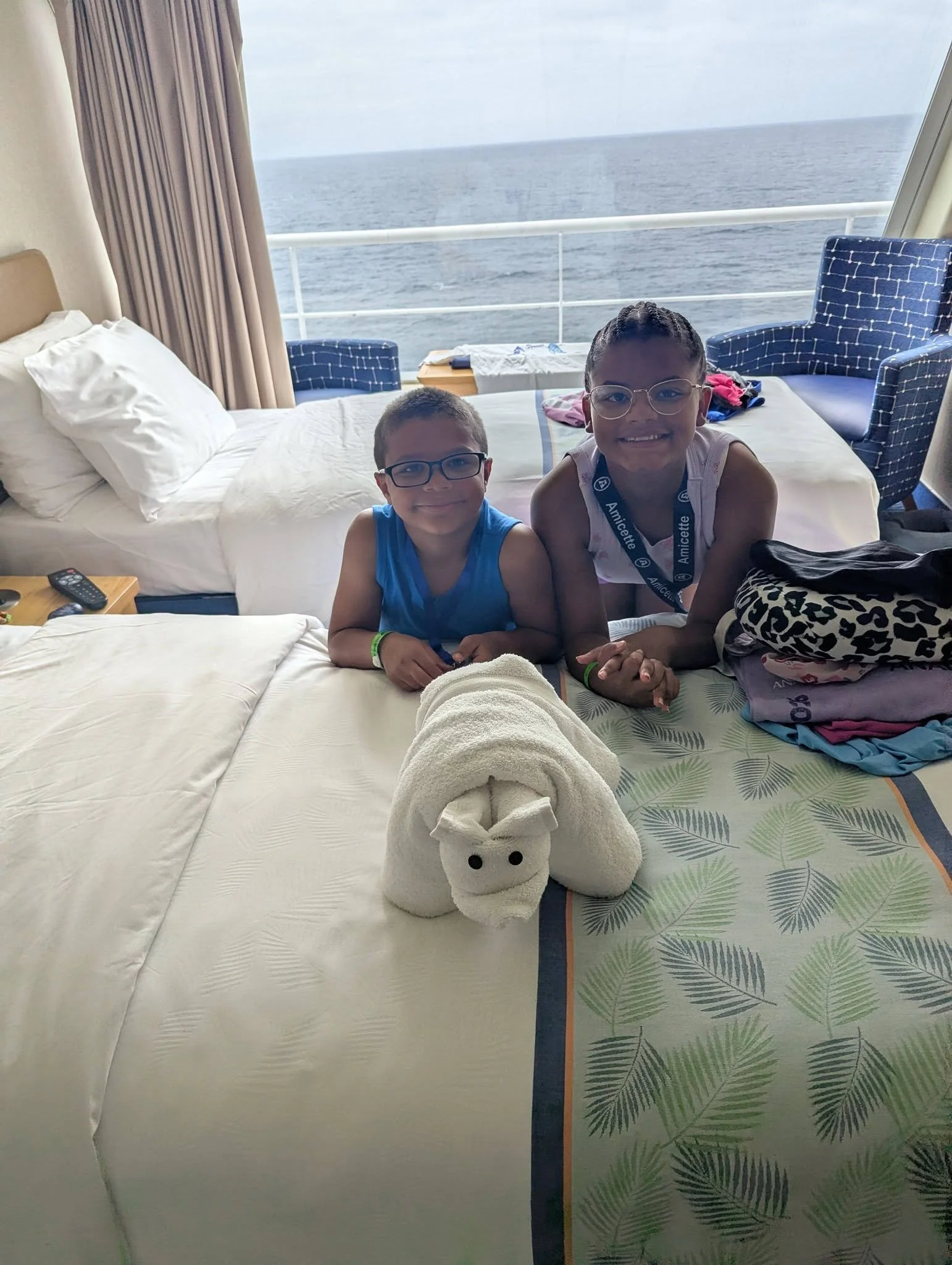 A hotel room with a view of the ocean through a large window. Two children with glasses, a boy and a girl, are smiling at the camera. A towel folded into a bunny shape is in the foreground. The bed has white linens, and there are two blue chairs and 