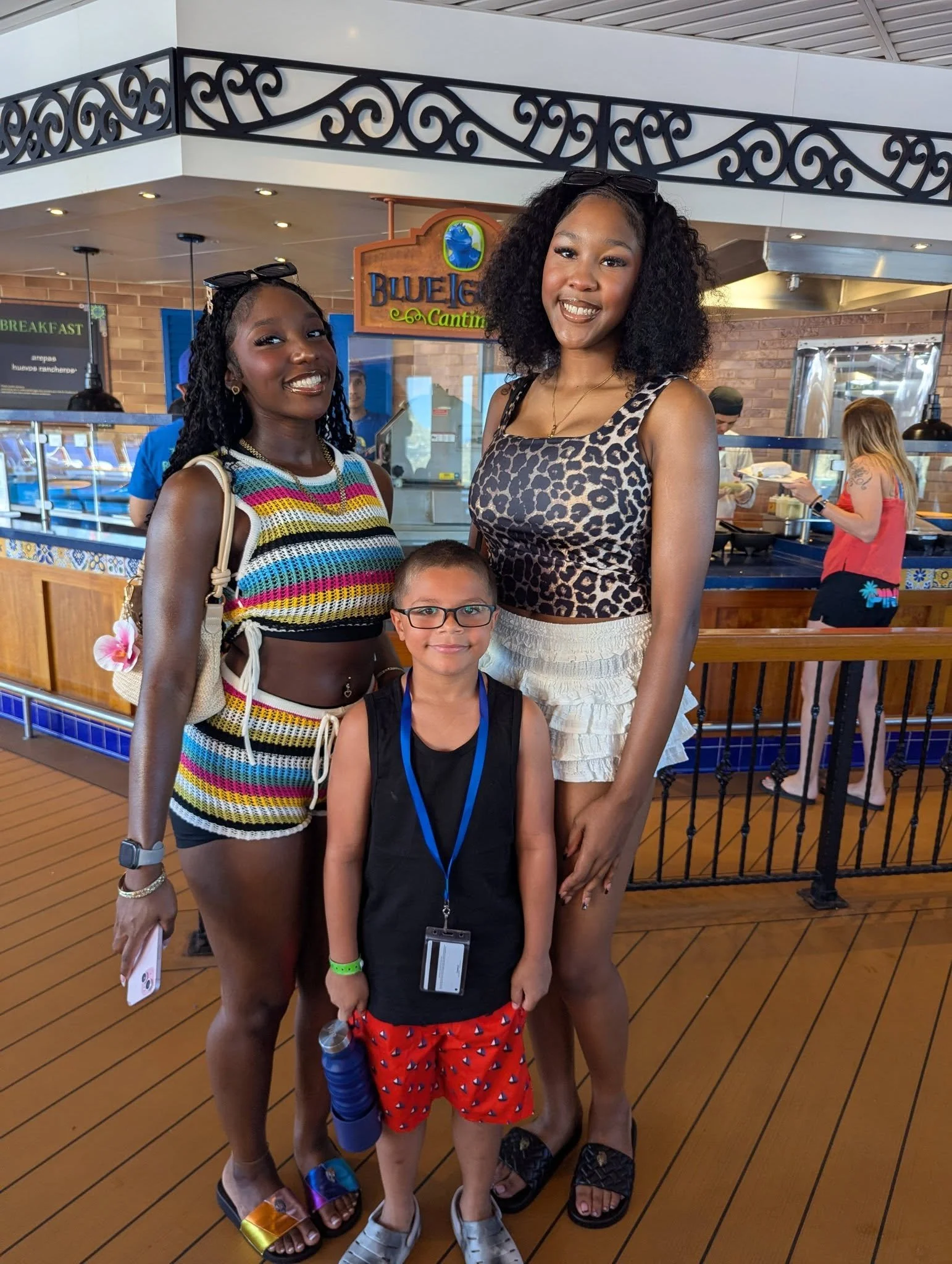 Two women and a boy standing together in a restaurant, with the women smiling and the boy looking at the camera. The women are wearing fashionable summer clothing and sandals, and the boy is wearing glasses, a black sleeveless shirt, and red shorts. 