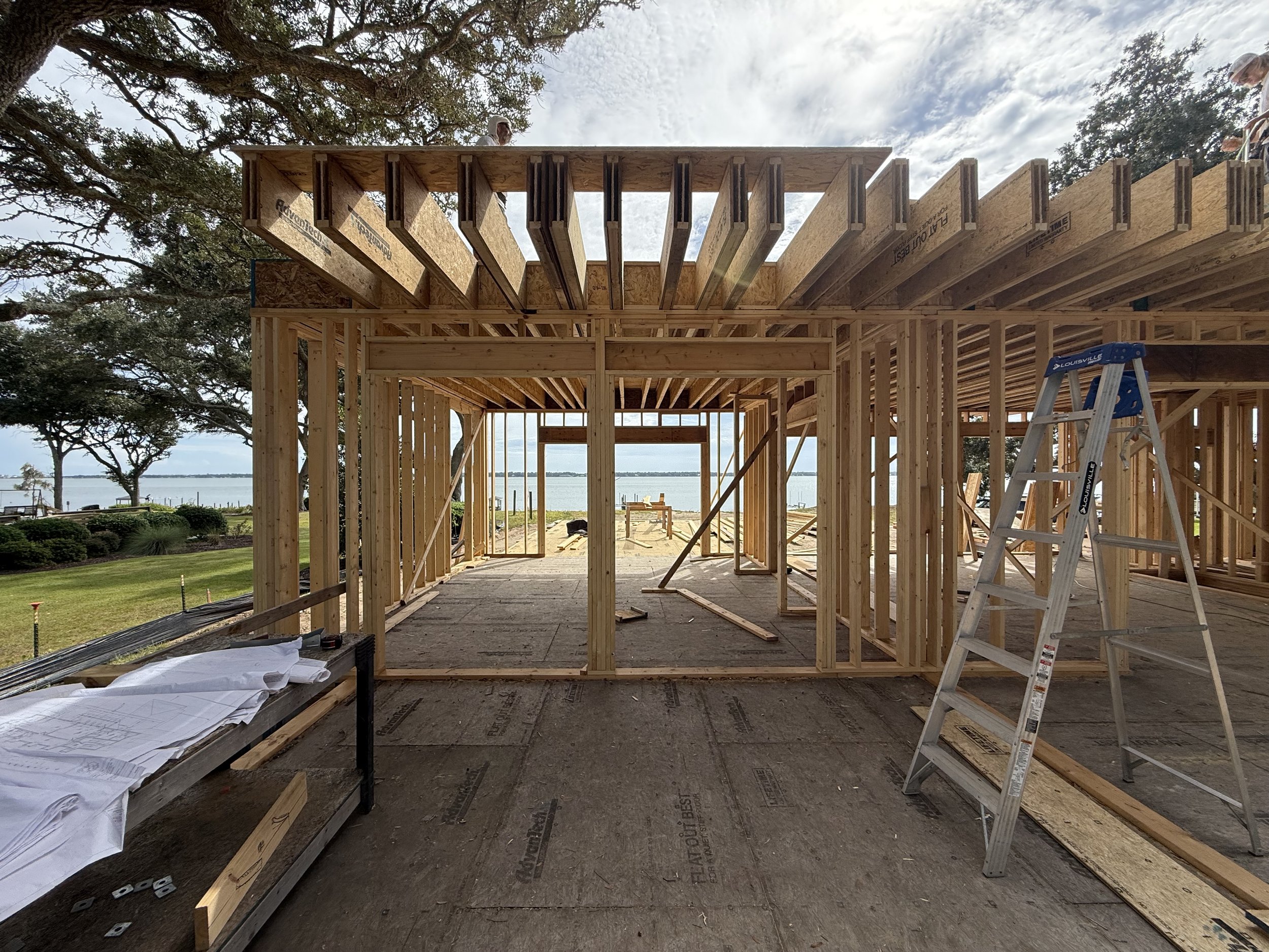 Construction site of a wooden house or building with framed walls, stairs, and a ladder, overlooking a body of water with trees in the background.